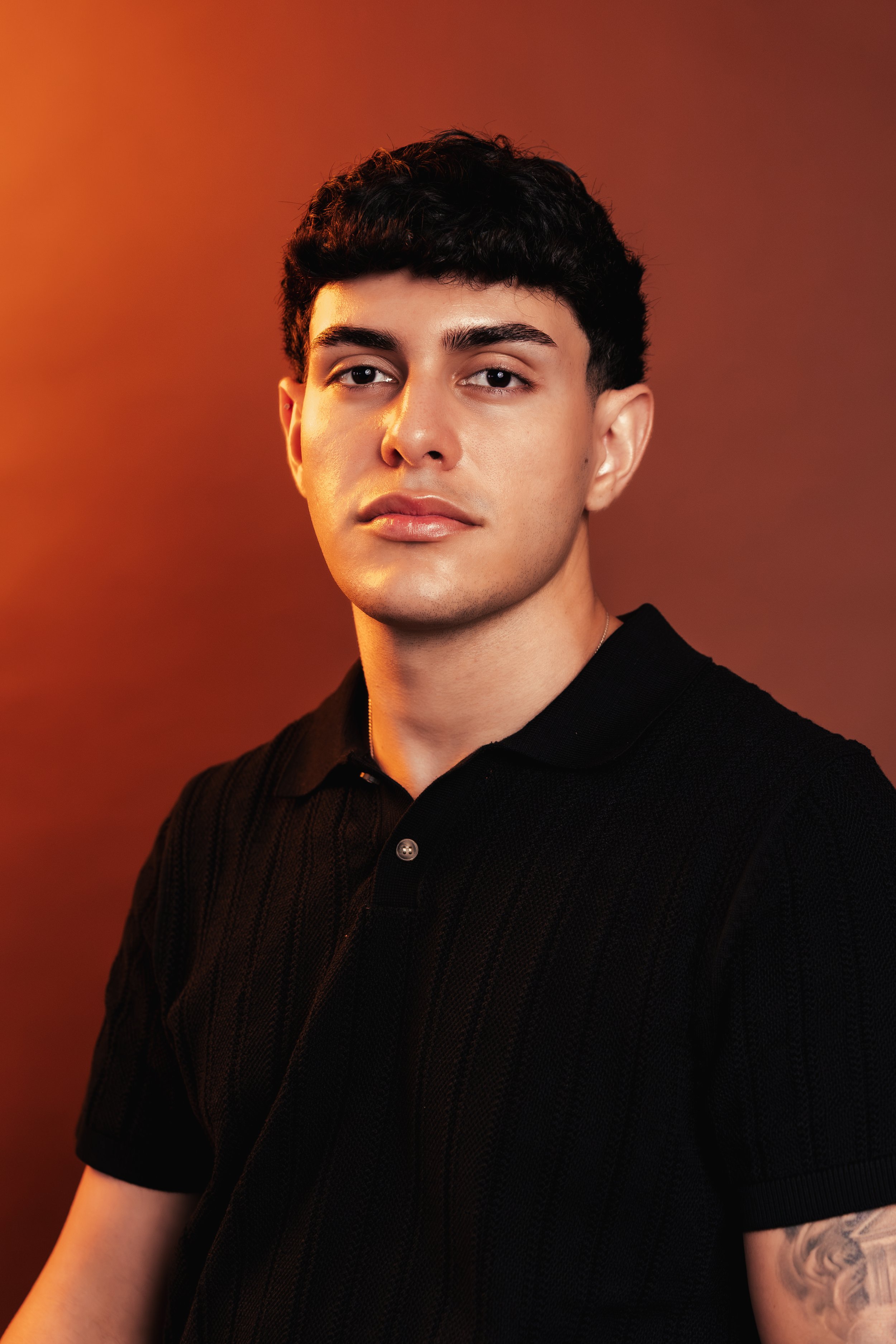 Young man with dark hair and a tattoo on his arm, wearing a black shirt, posing against a warm-toned background.
