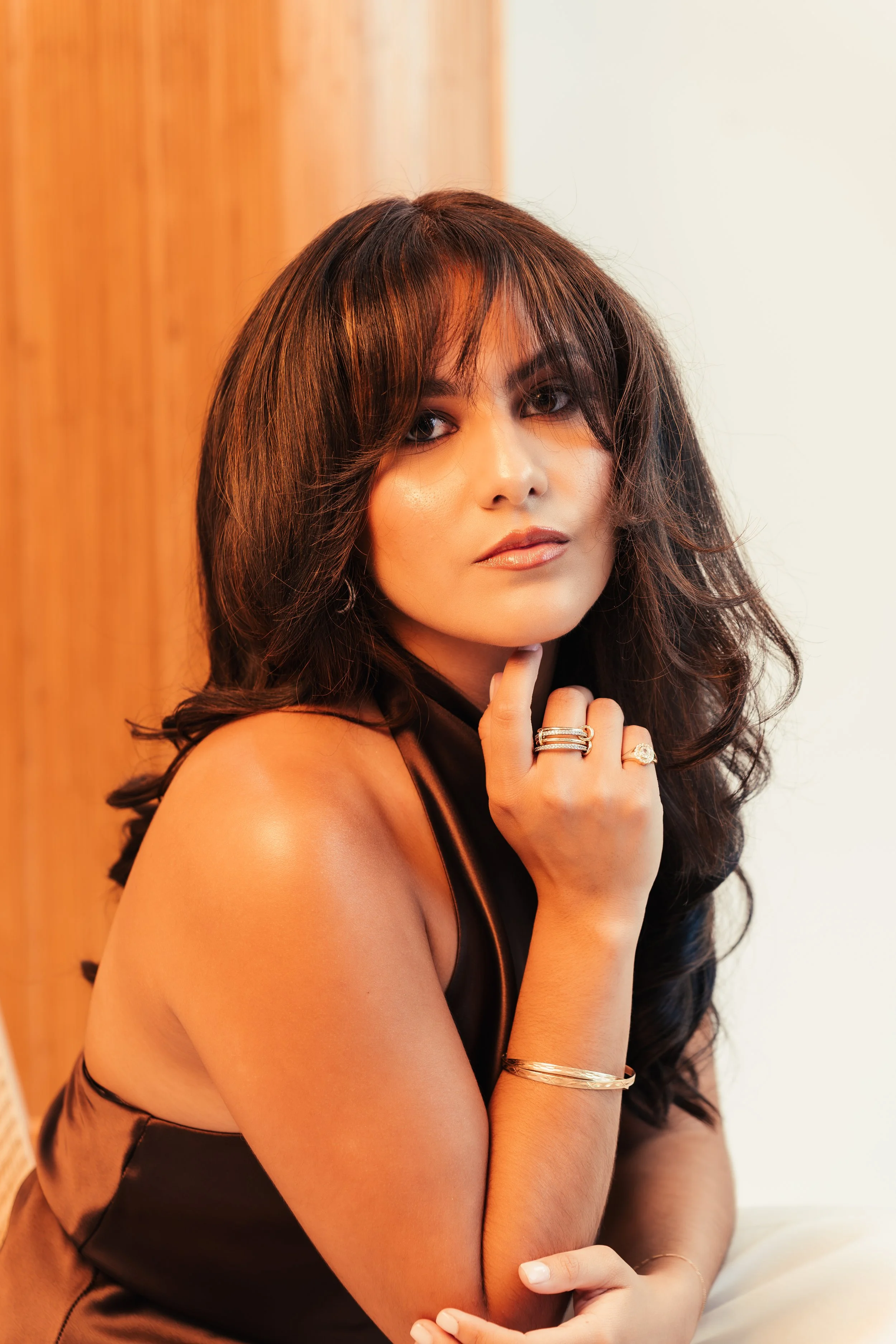 Portrait of a woman with dark, wavy hair, wearing gold jewelry, posing with her hand on her chin in front of a wooden and white background.