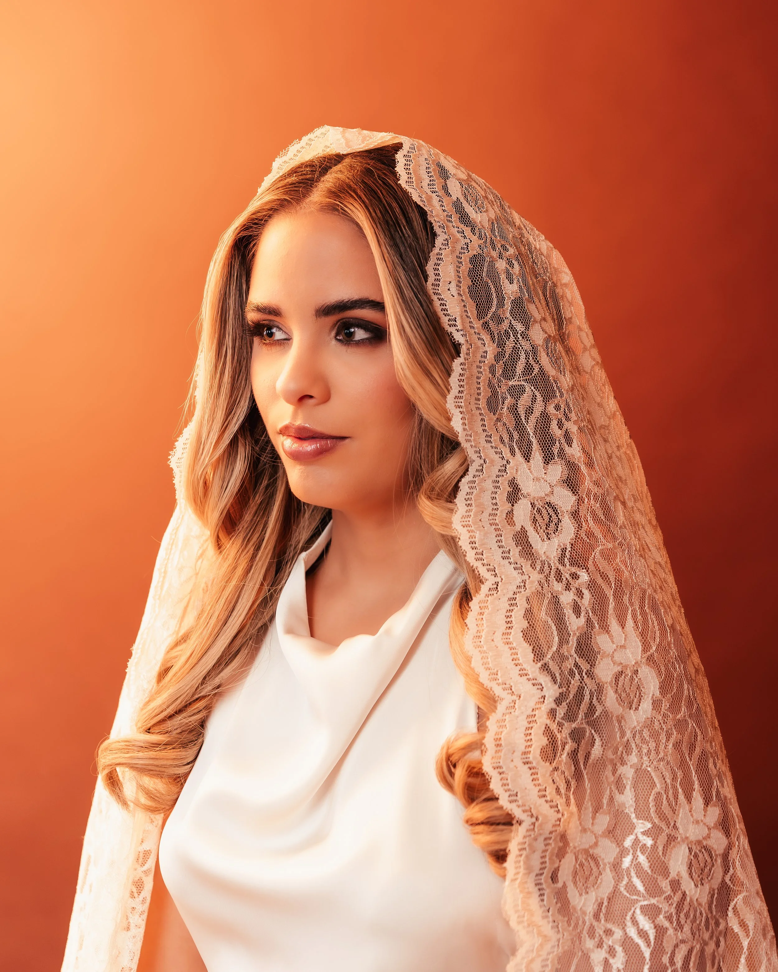 A woman with long, wavy hair, wearing a white silky top and a lace veil over her head, against an orange gradient background.