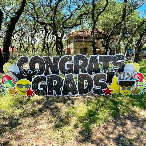 Decorative sign that says 'Congrats Grads' with balloons, stars, and sunglasses icons, in a park with trees and a building in the background.
