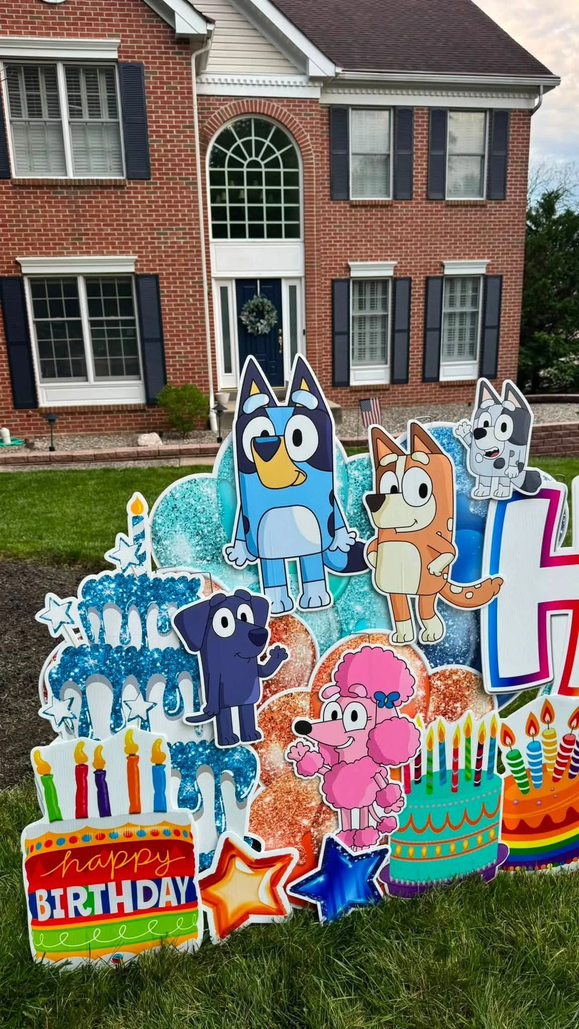 Colorful birthday yard decorations with cartoon dogs, birthday candles, stars, and a 'happy birthday' sign in front of a brick house with white-framed windows.