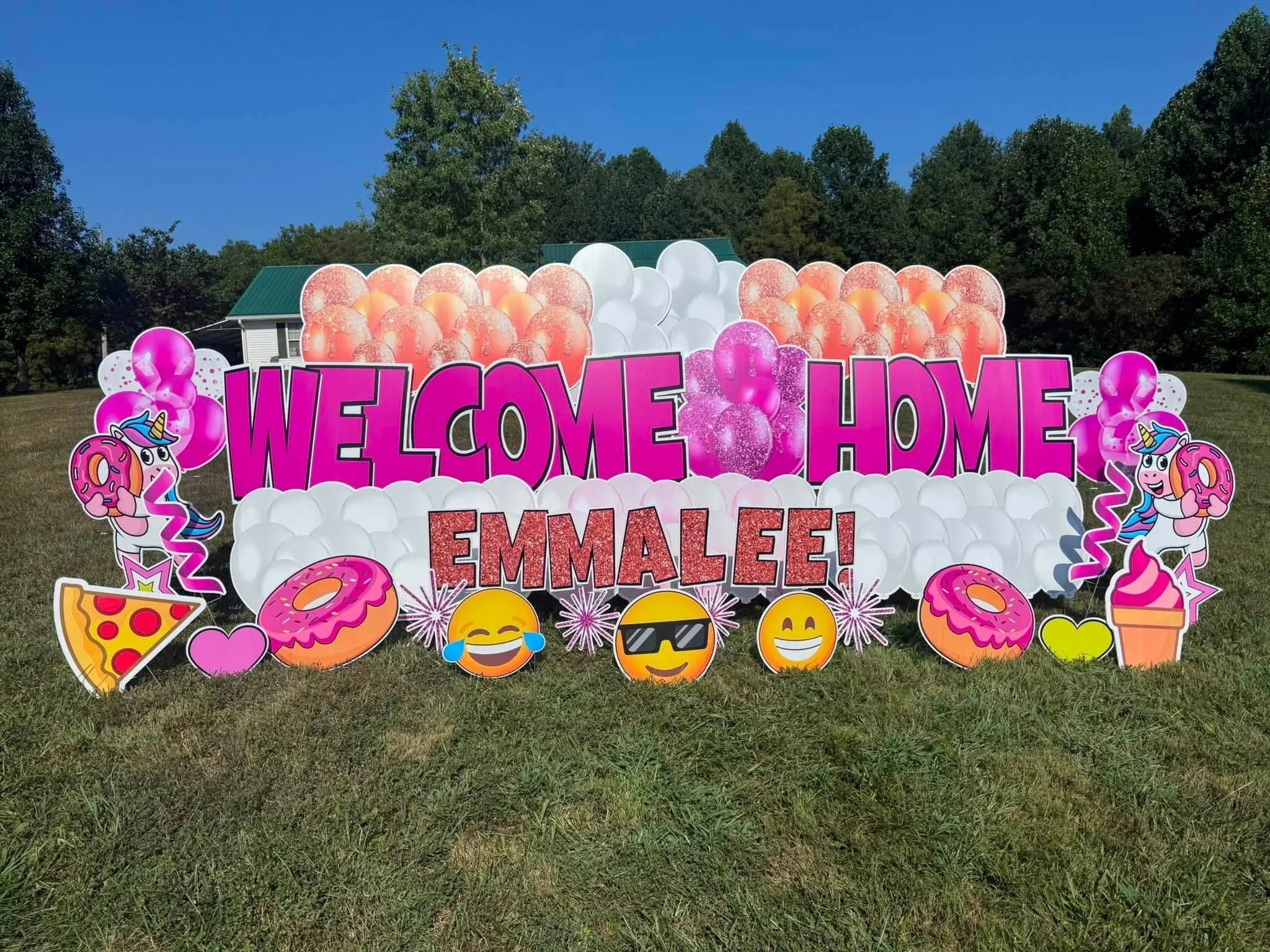 Decorative sign with pink, orange, and white balloons, featuring the message "Welcome Home Emmalee!" and surrounded by cartoonish emoji and ice cream graphic illustrations, set outdoors on grass with trees and a house in the background.