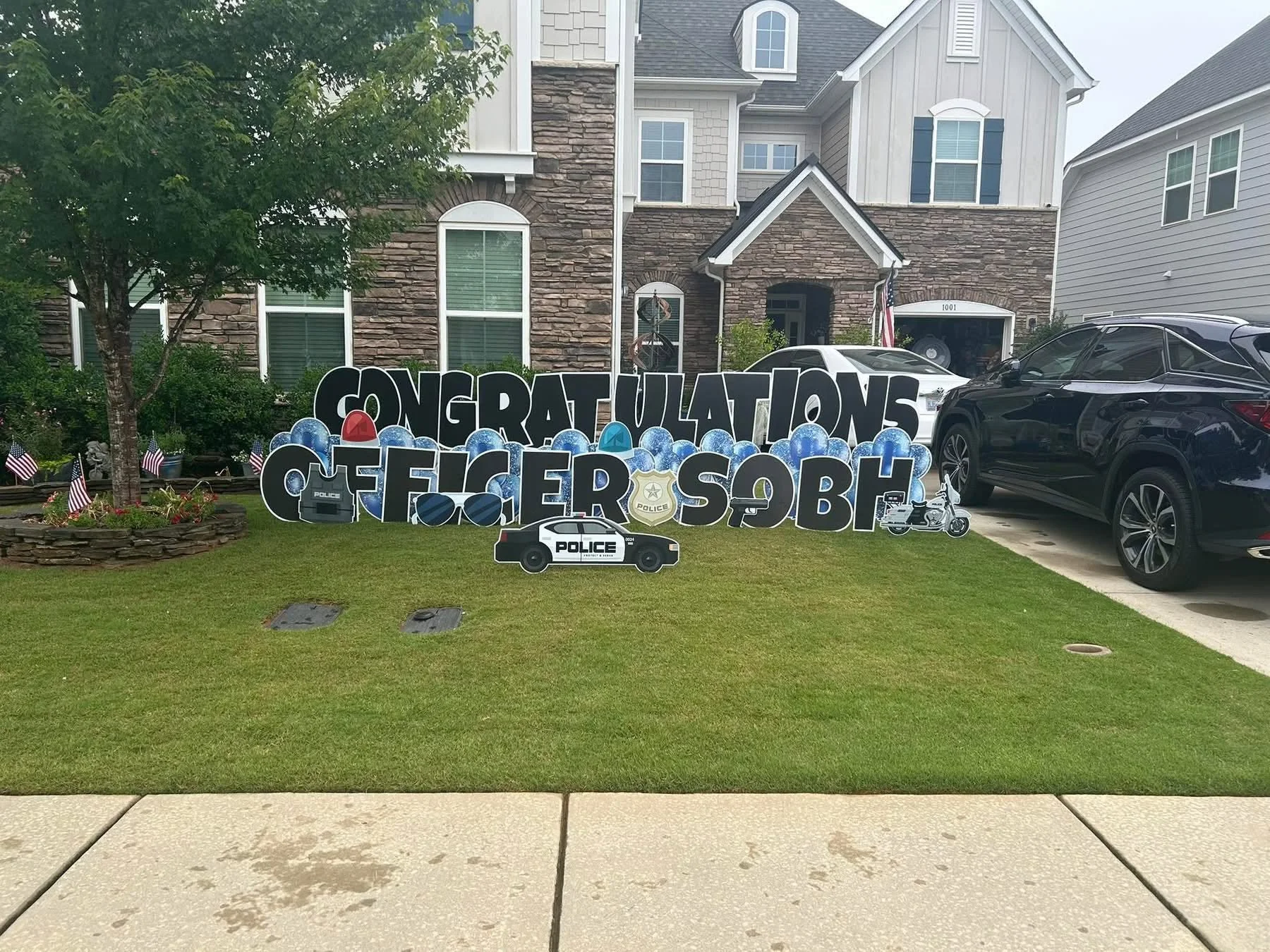 Front yard of a house decorated with a large sign that says "Congratulations Officer SOB". The sign includes police-themed images like badges, a police car, and a motorcycle. Small American flags are planted in the flower bed, and there are three cars parked in the driveway. The house features a brick and siding exterior with white window trims and a small front porch.