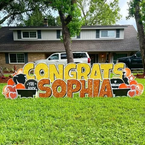 Colorful graduation sign with 'Congrats Sophia' on a grassy lawn in front of a house, decorated with graduation-themed icons like caps, diplomas, and hearts.