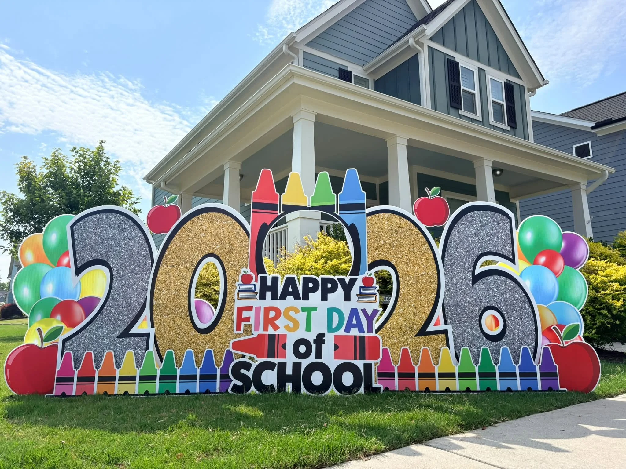 Colorful first day of school sign with numbers, balloons, and pencils in front of a two-story house with a porch, under a partly cloudy sky.