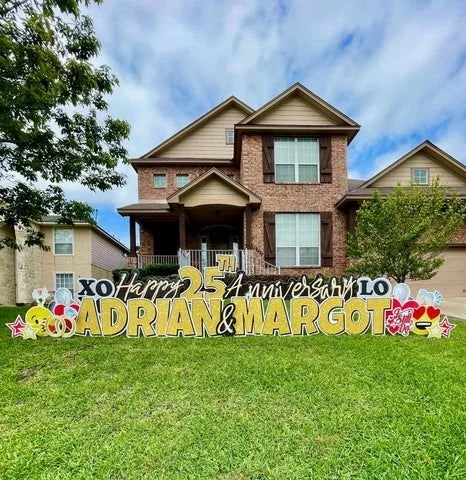 Front yard of a two-story house with a large sign celebrating Adrian and Margot's 25th wedding anniversary, decorated with colorful symbols and text.
