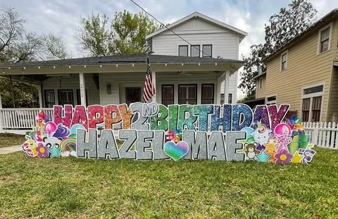 Colorful birthday yard sign with the message "HAPPY 2ND BIRTHDAY HAZE MAE" decorated with flowers, hearts, and cartoon characters on a lawn in front of a house.