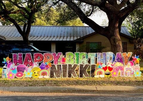 Colorful birthday signs reading 'Happy Birthday Nikkie' with floral and emoji decorations outside next to a tree and a house.