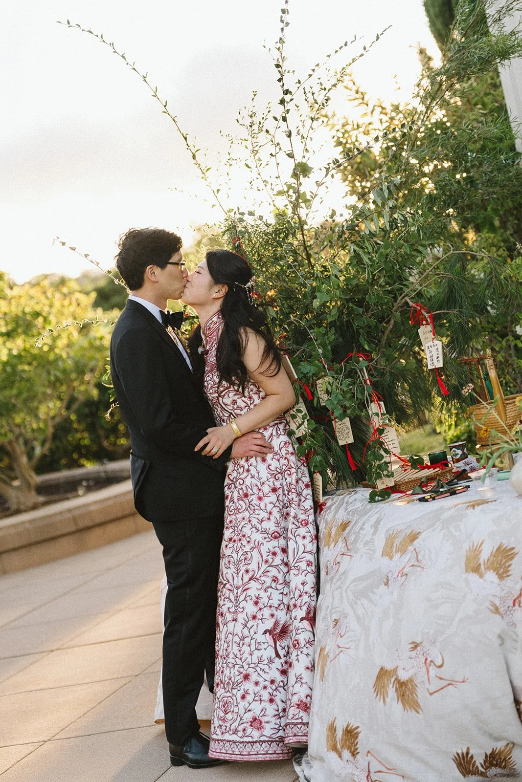 A couple in wedding attire kissing at an outdoor wedding reception, with greenery and wedding decorations in the background.