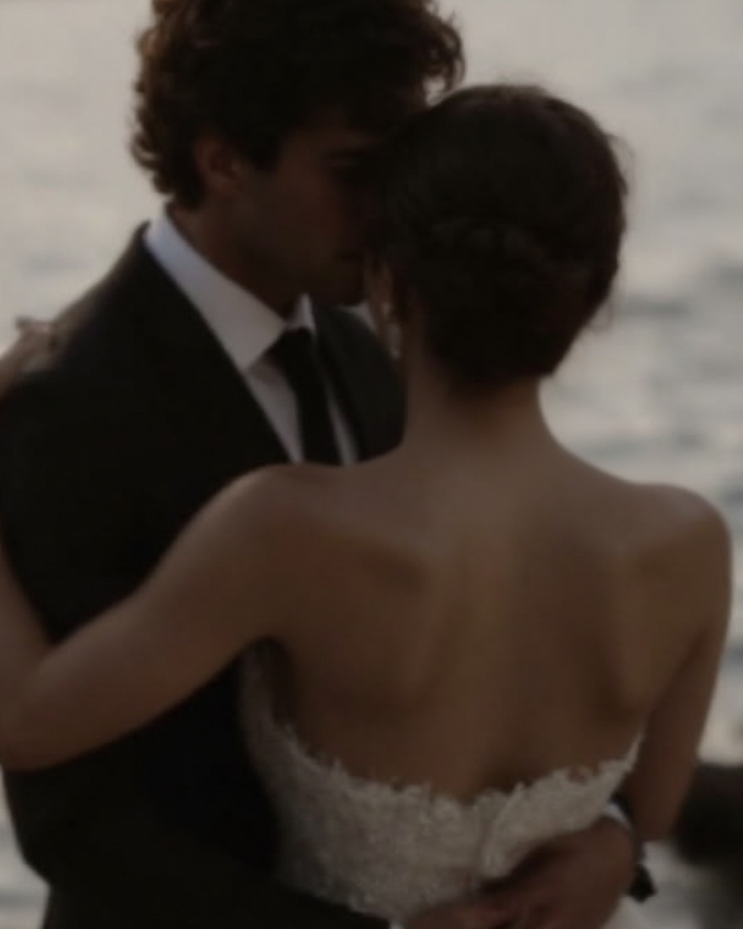 A man and woman in formal attire close together, with their foreheads touching in an intimate moment, against a light-colored background.