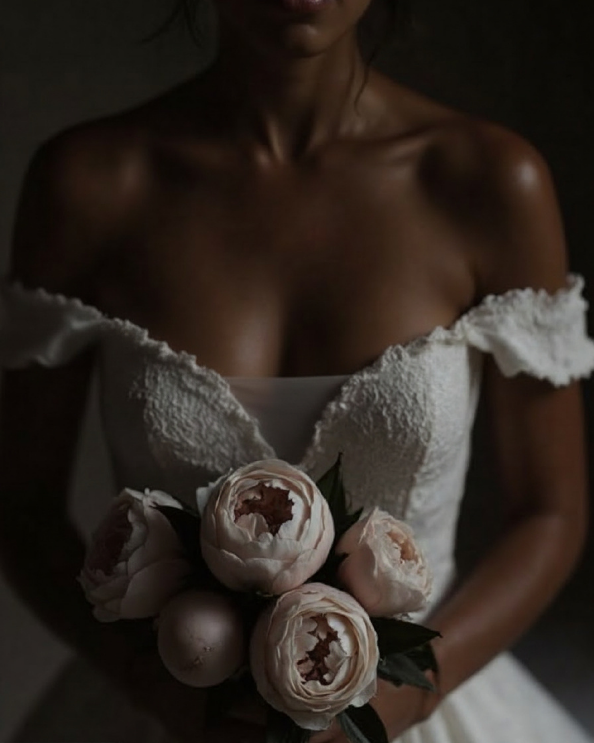 A woman with dark skin and dark hair, partly visible, holding a bouquet of pink and white roses wrapped in white paper, against a dark background.