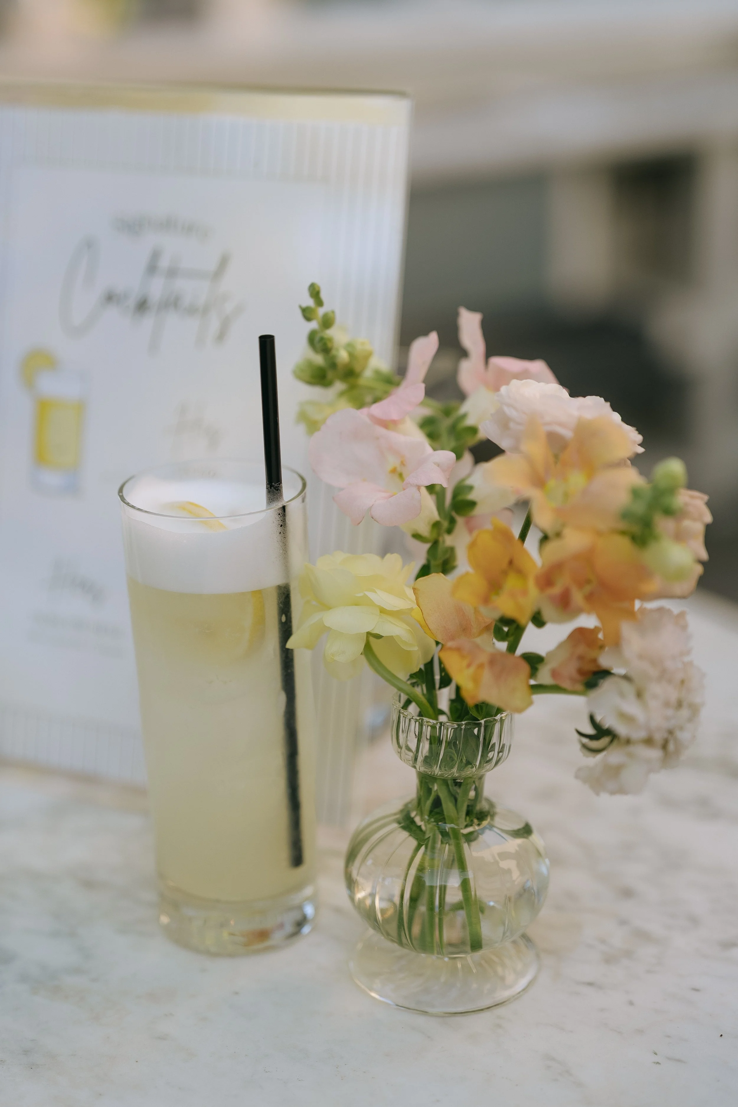 A clear glass vase filled with pastel-colored flowers, placed on a white surface, with a cocktail menu and a tall drink with a black straw visible in the background.