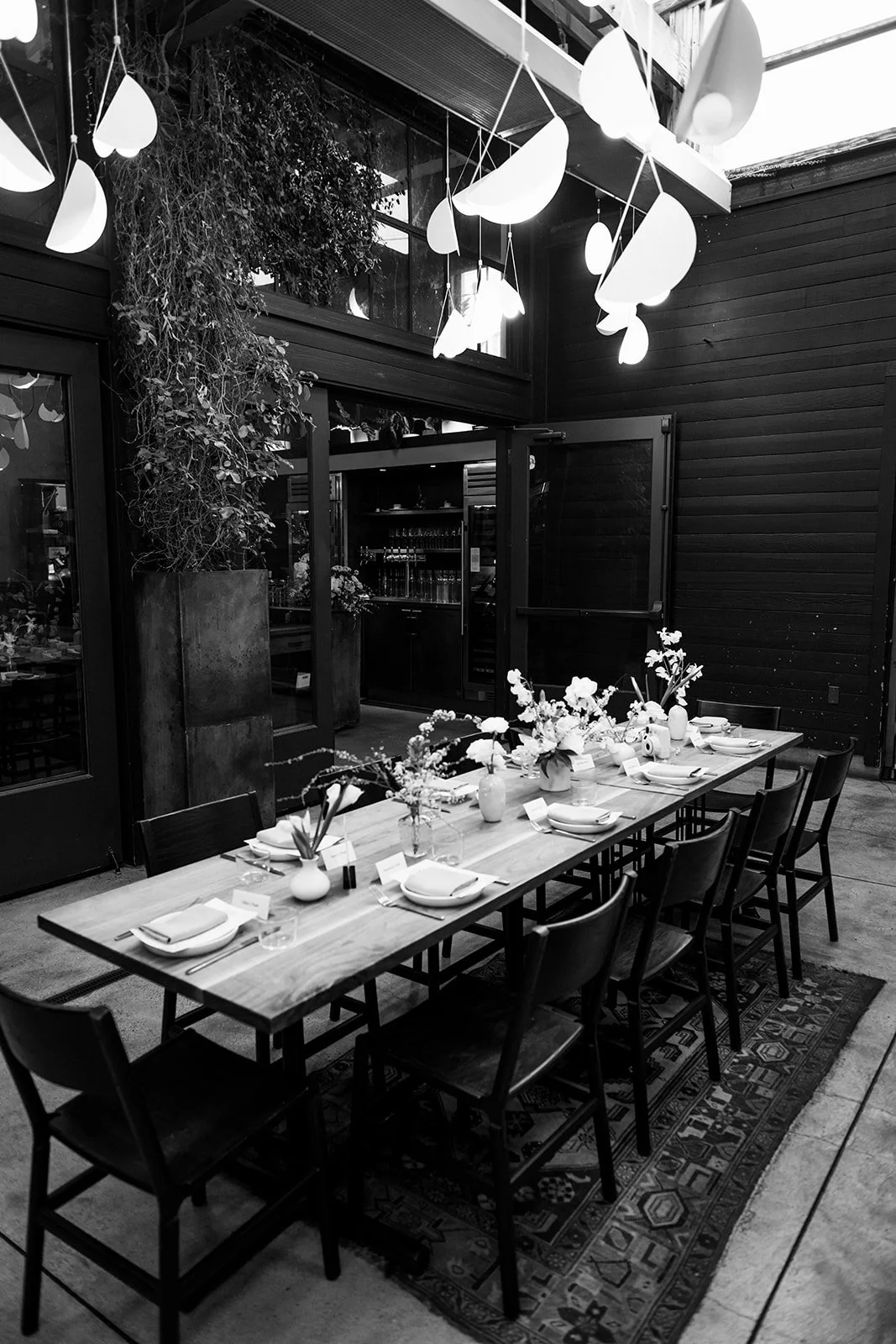 A dining table set for a meal with plates, glasses, napkins, and floral centerpieces in a modern restaurant with dark walls, hanging leaf-shaped decorations, and large windows.