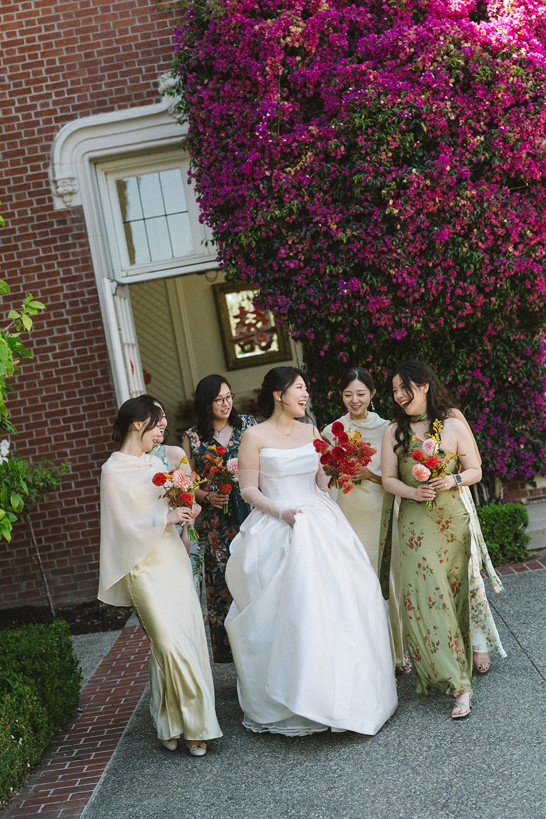 A bride and four women, possibly bridesmaids or friends, standing outside a house with a large pink flowering bush in the background. They are smiling and holding bouquets of flowers.