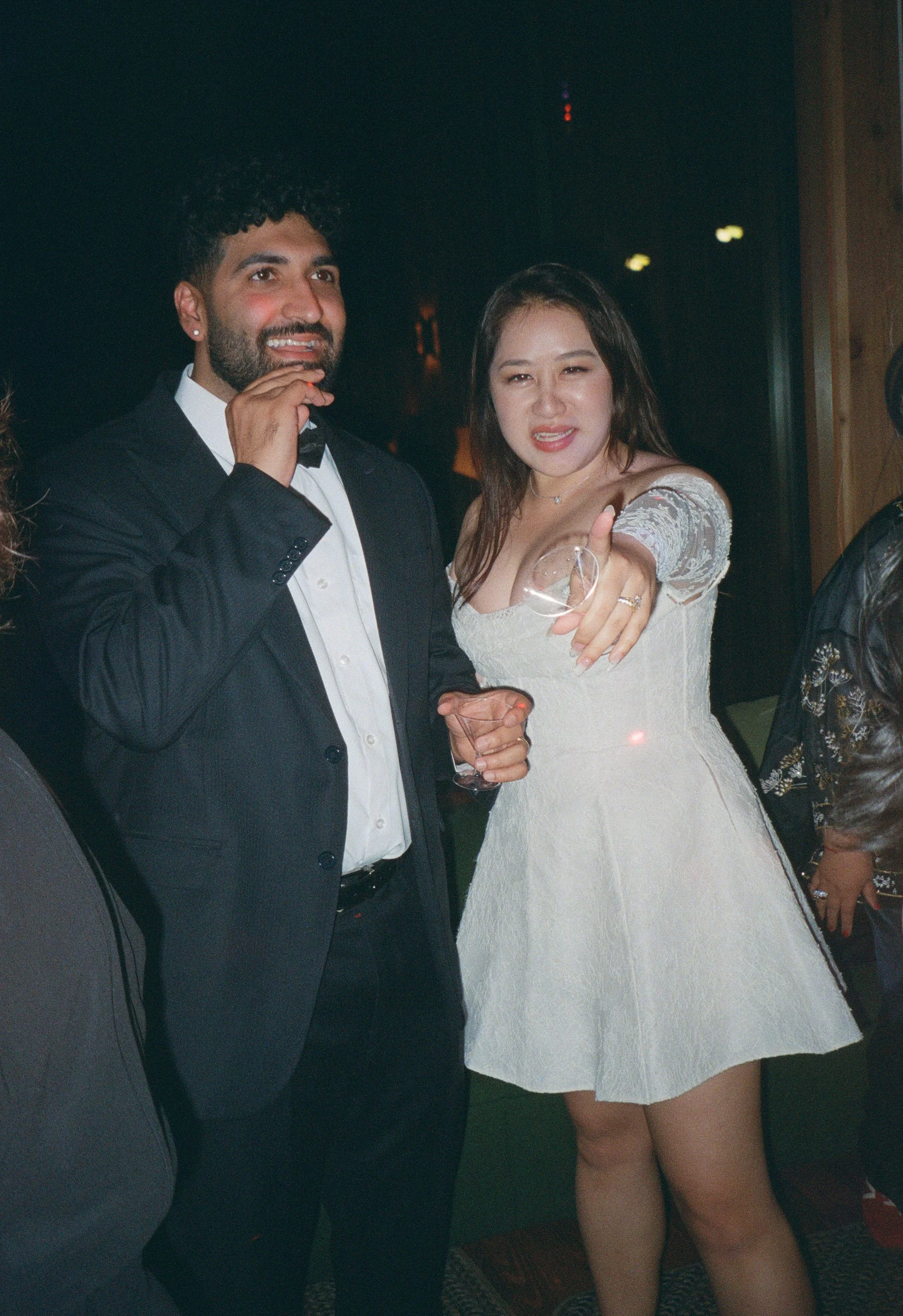Two people, a man in a tuxedo and a woman in a white dress, are at a celebration or party, smiling and holding drinks.