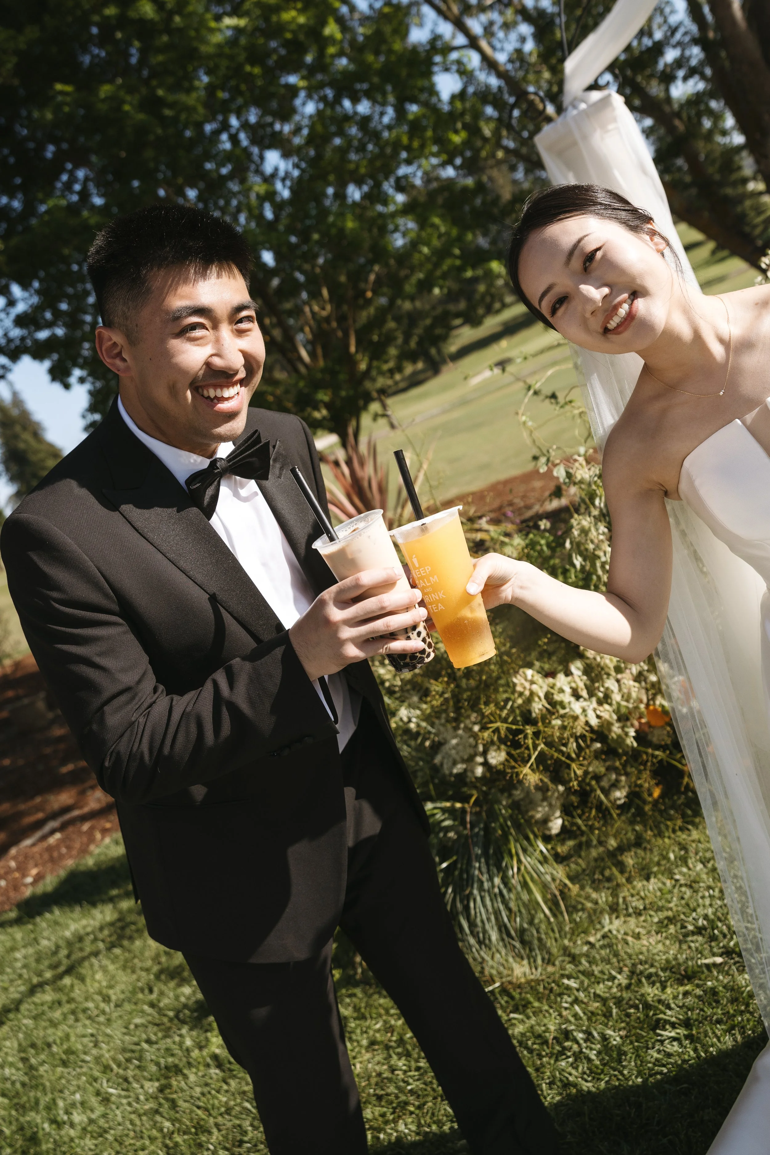 A man in a black tuxedo and a woman in a white wedding dress are smiling and holding drinks at an outdoor wedding celebration. They are standing on grass with trees and a white decorative element in the background.