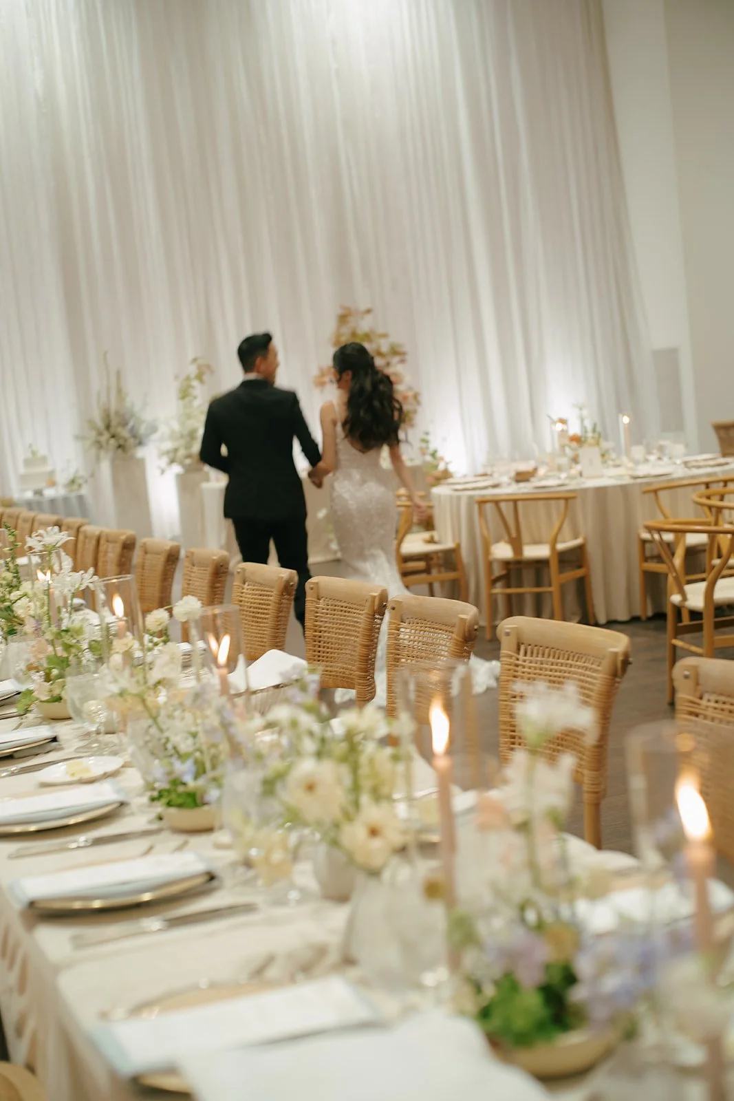 A bride and groom holding hands, walking in a decorated wedding reception hall with tables, floral arrangements, and candle lighting.