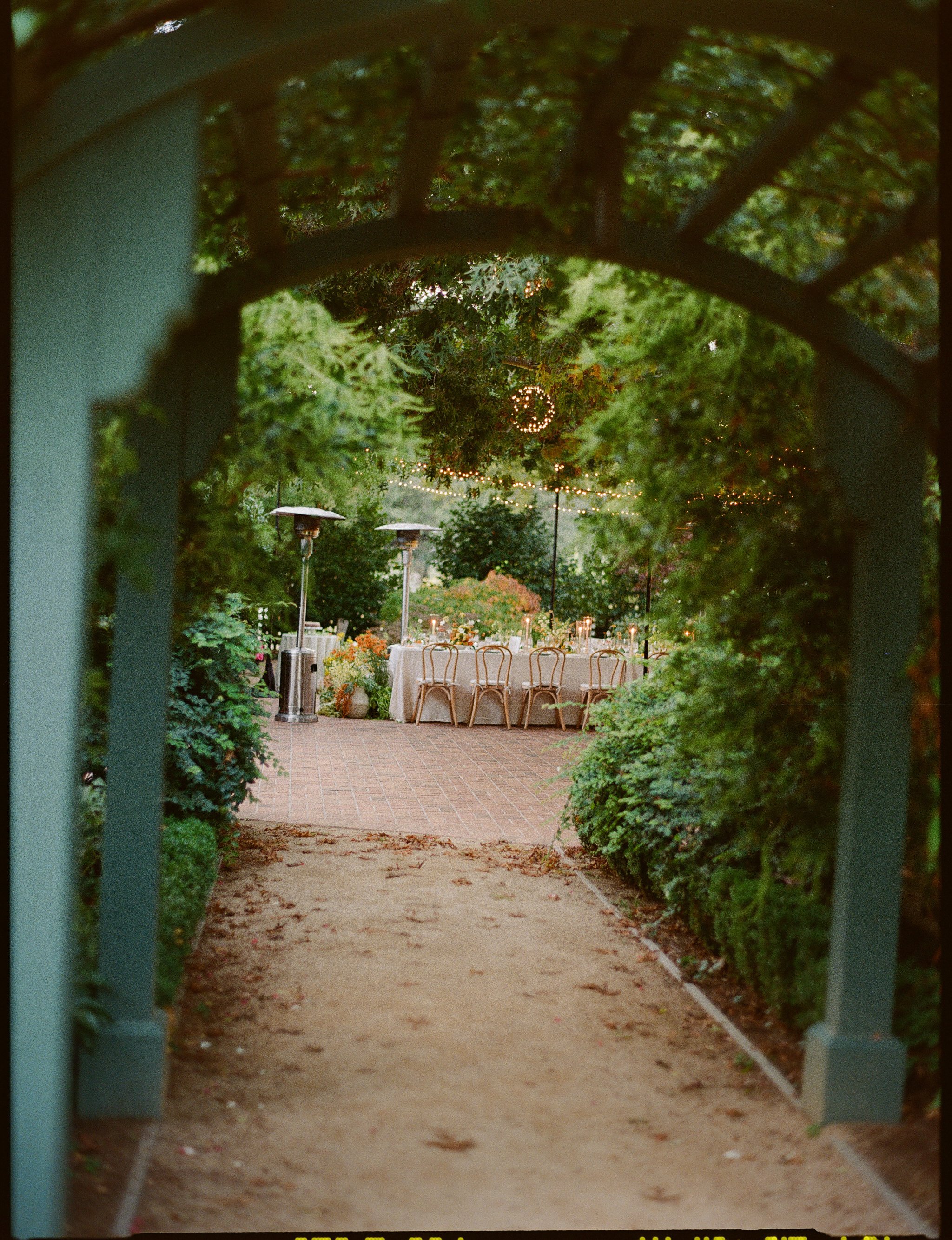 View through a green garden arbor to a patio with a long table, chairs, string lights, and outdoor heaters.