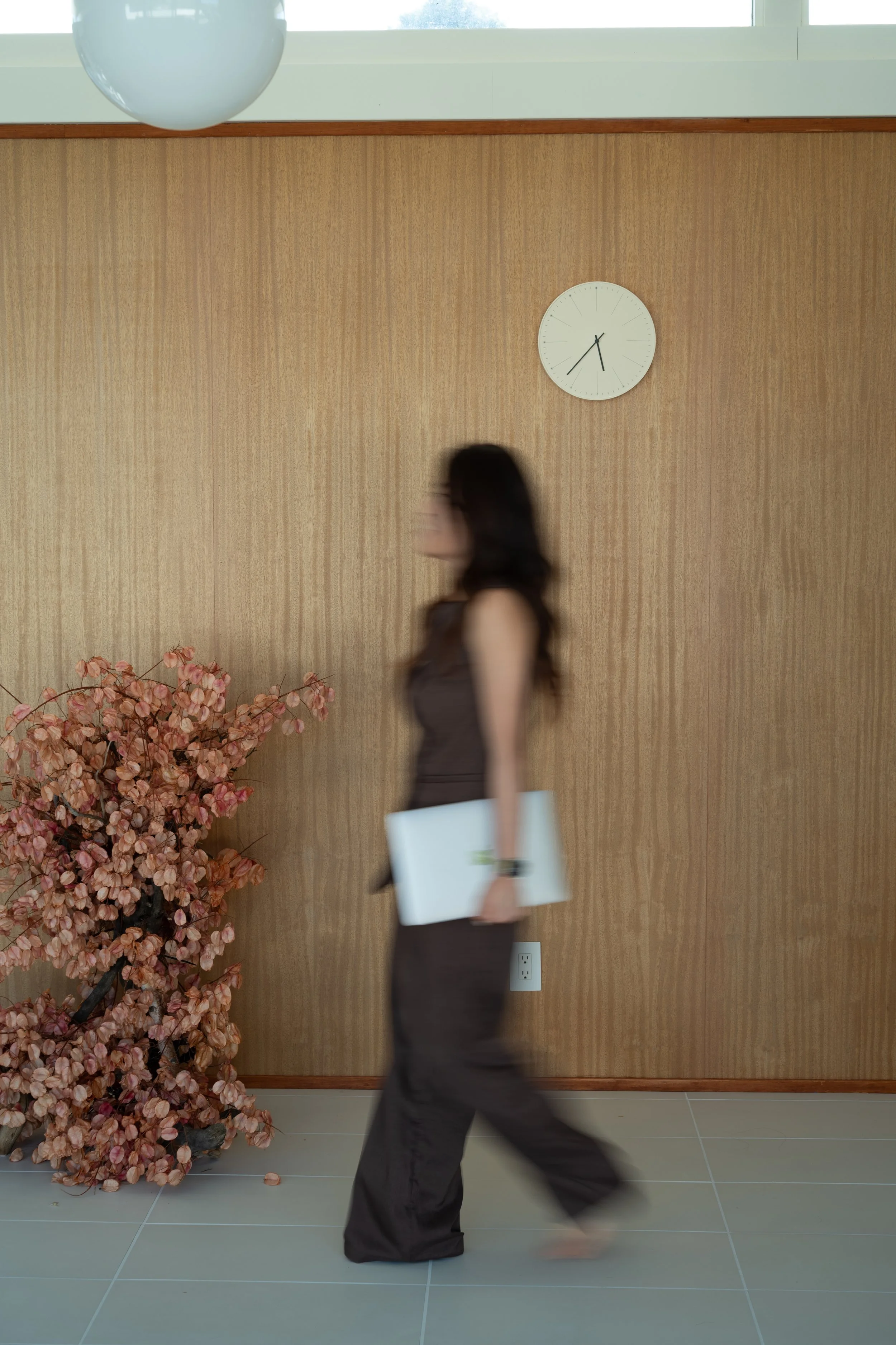 An indoor scene with a woman walking, holding a white folder or tablet, in front of a beige wooden wall with a round white wall clock showing 12:24. To the left, pinkish-orange flowers are in a large pot, and there is an electrical outlet at the bottom of the wall. A large round light fixture hangs from the ceiling.