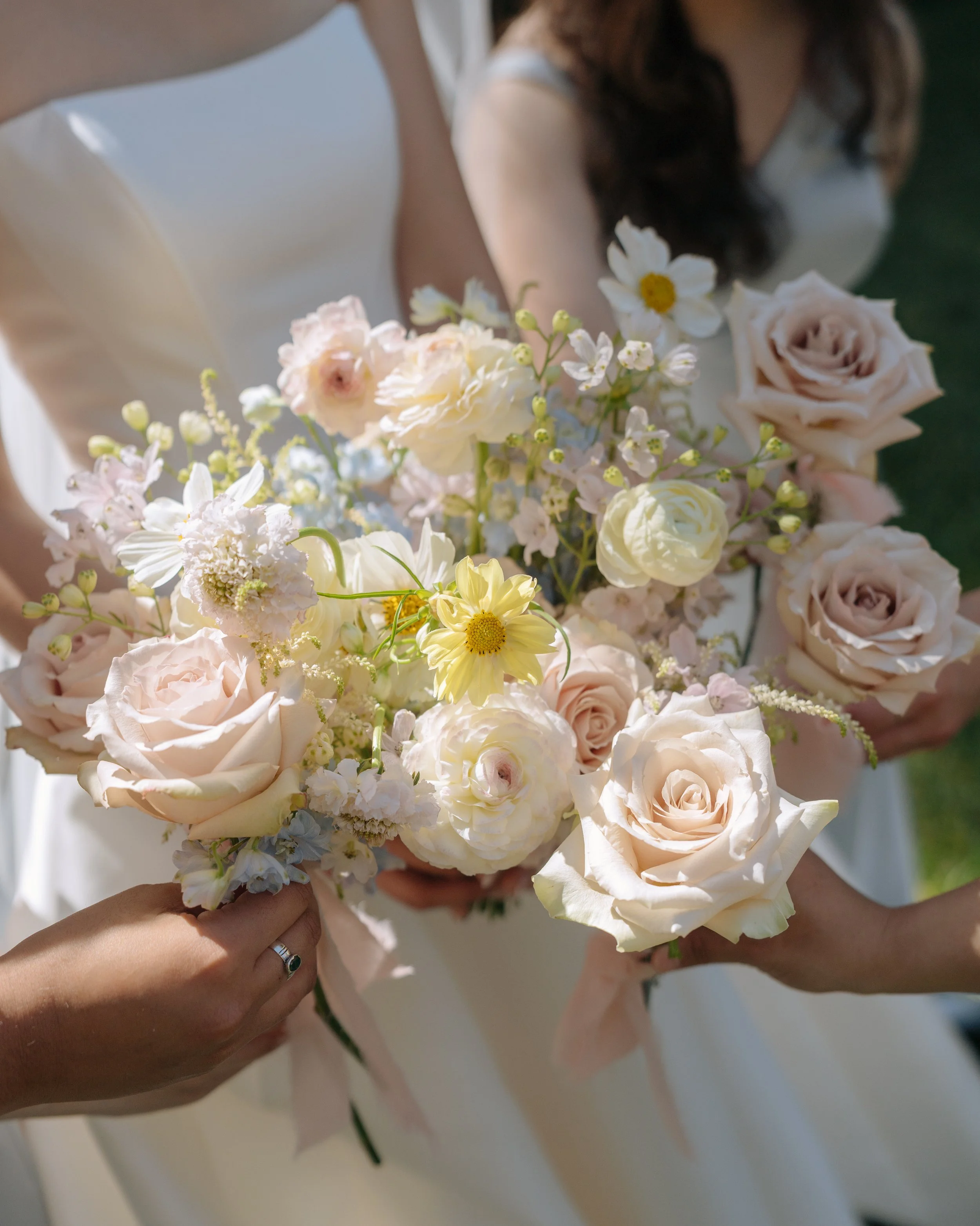 A bride holding a bouquet of white and pale pink flowers, including roses and small daisies, with her hand displaying a ring.