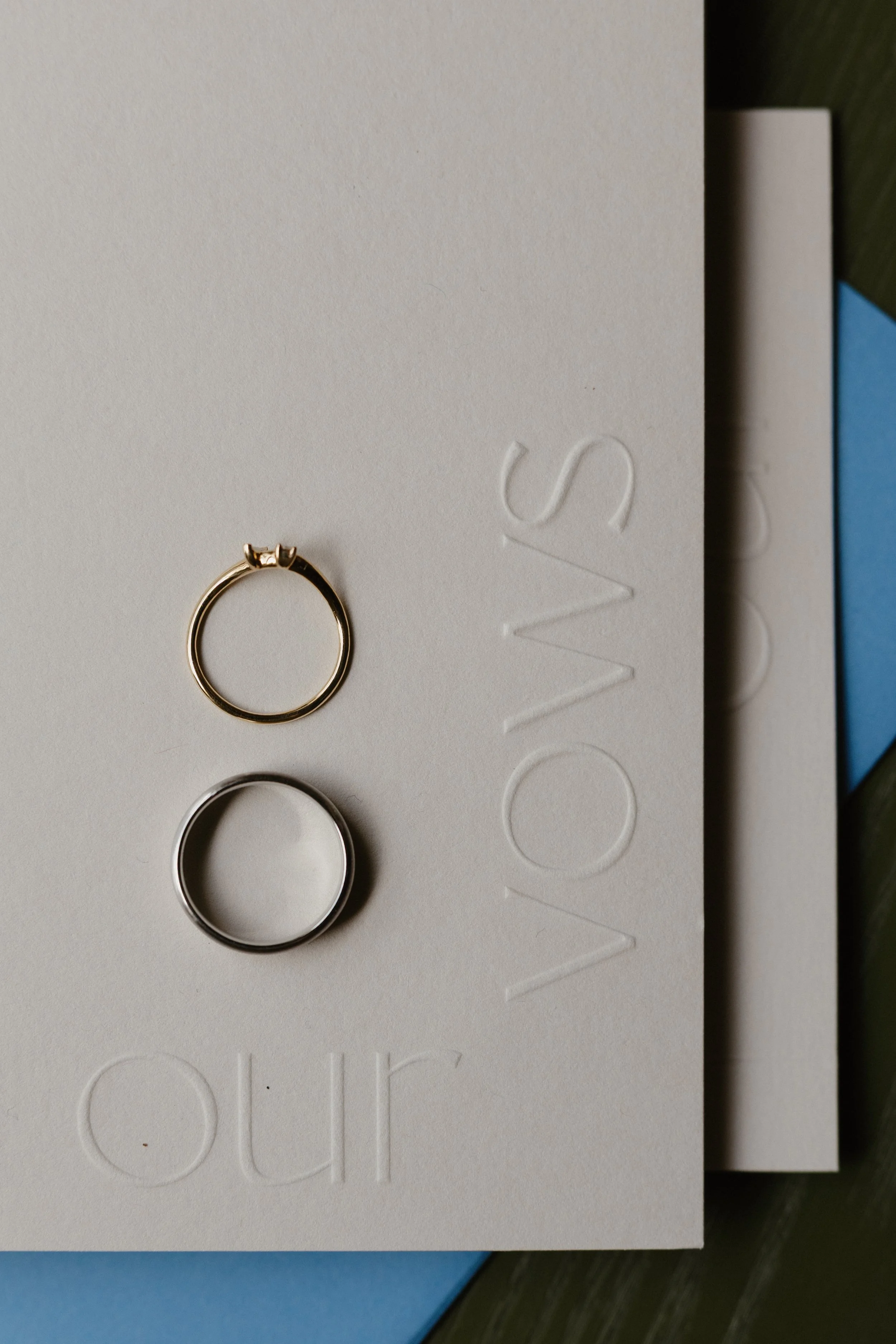 Close-up of three rings, two silver and one gold, placed on white embossed paper with the word 'VOW' visible. The paper appears to be part of a card or invitation.