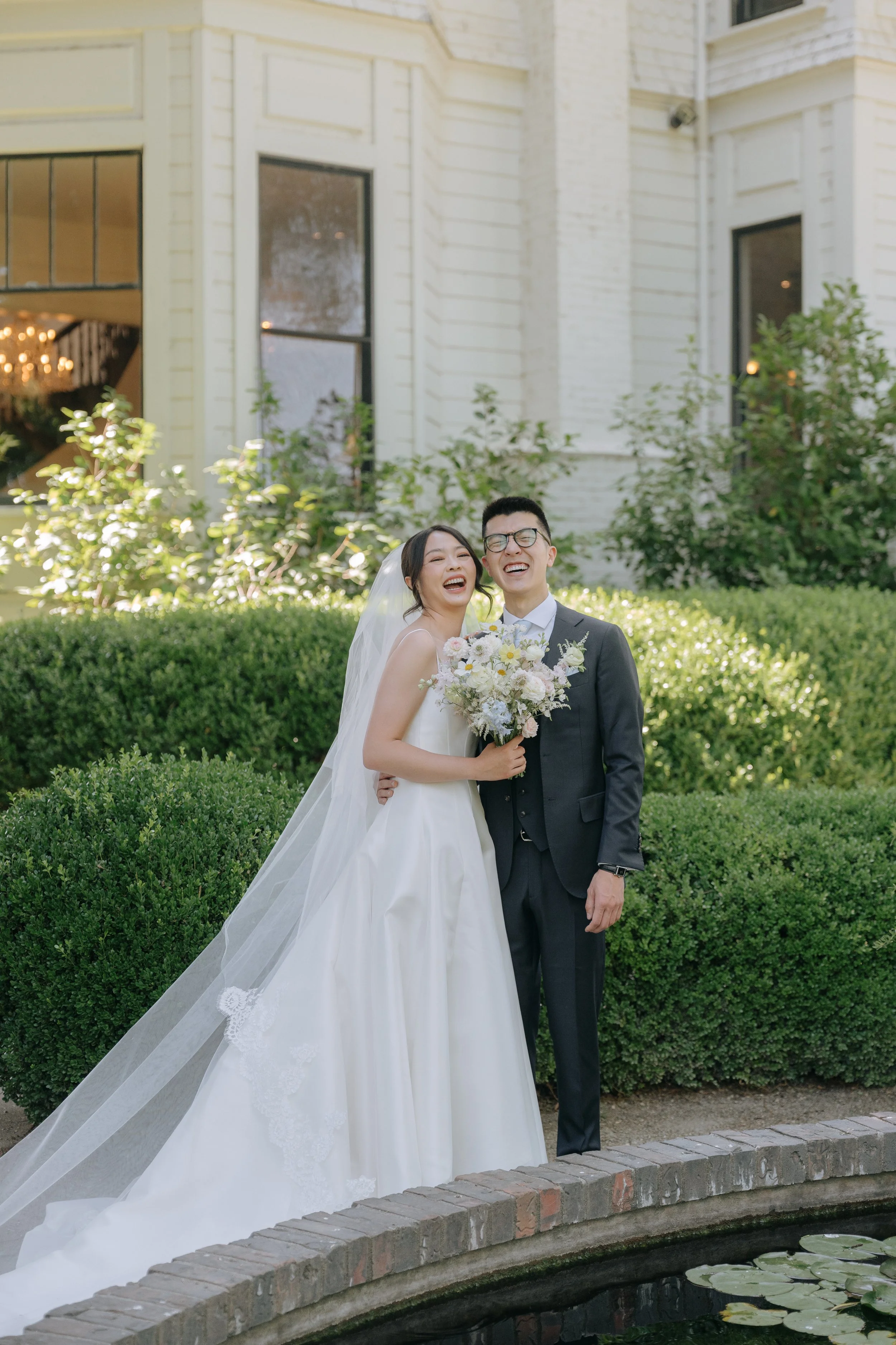A bride and groom walking together outdoors, smiling, with a white brick building and bushes in the background, near a small pond with lily pads.