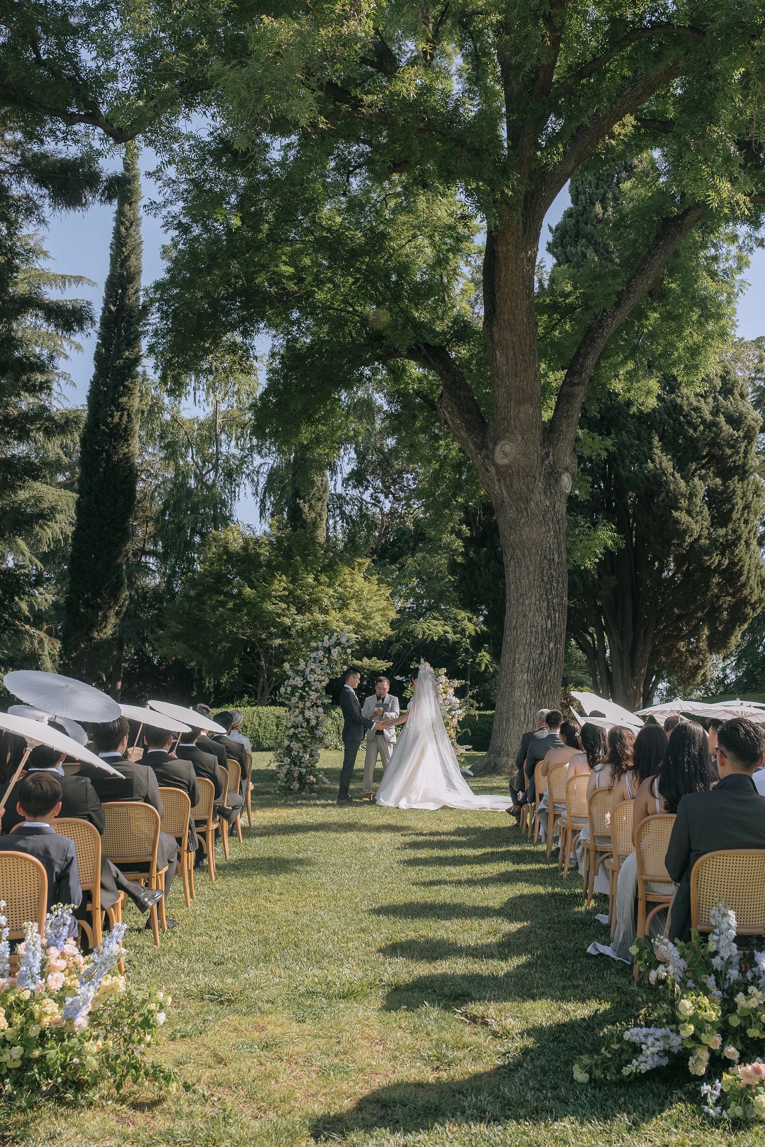 A outdoor wedding ceremony set in a garden with large trees, guests seated on chairs holding umbrellas, and a bride and groom standing at the altar with officiants. Flower arrangements decorate the aisle.