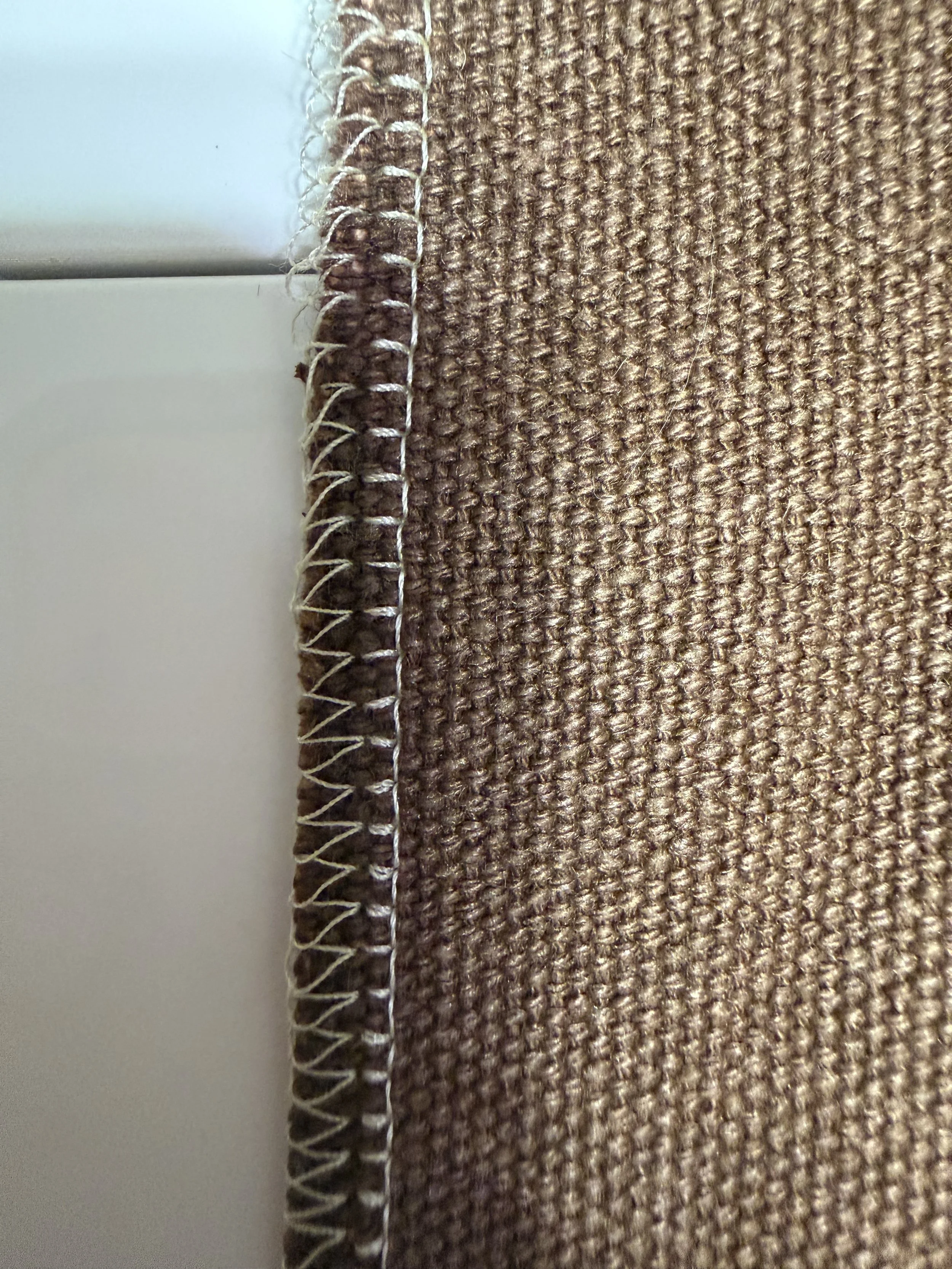 Close-up of a brown woven fabric with a white and brown striped thread along the edge, against a white background.