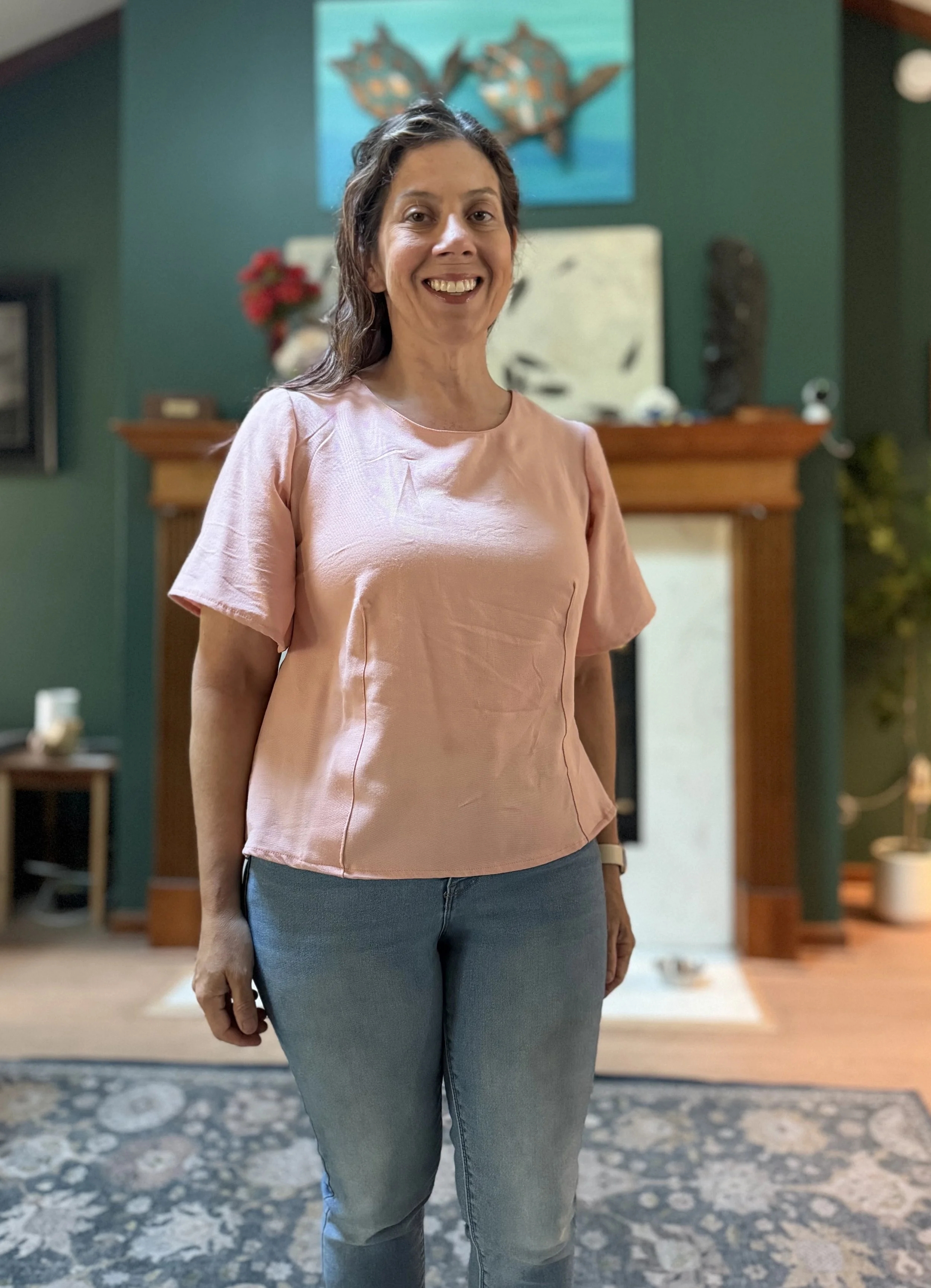 A woman standing inside a house, smiling at the camera, wearing a pink shirt and jeans, with a living room setting in the background.