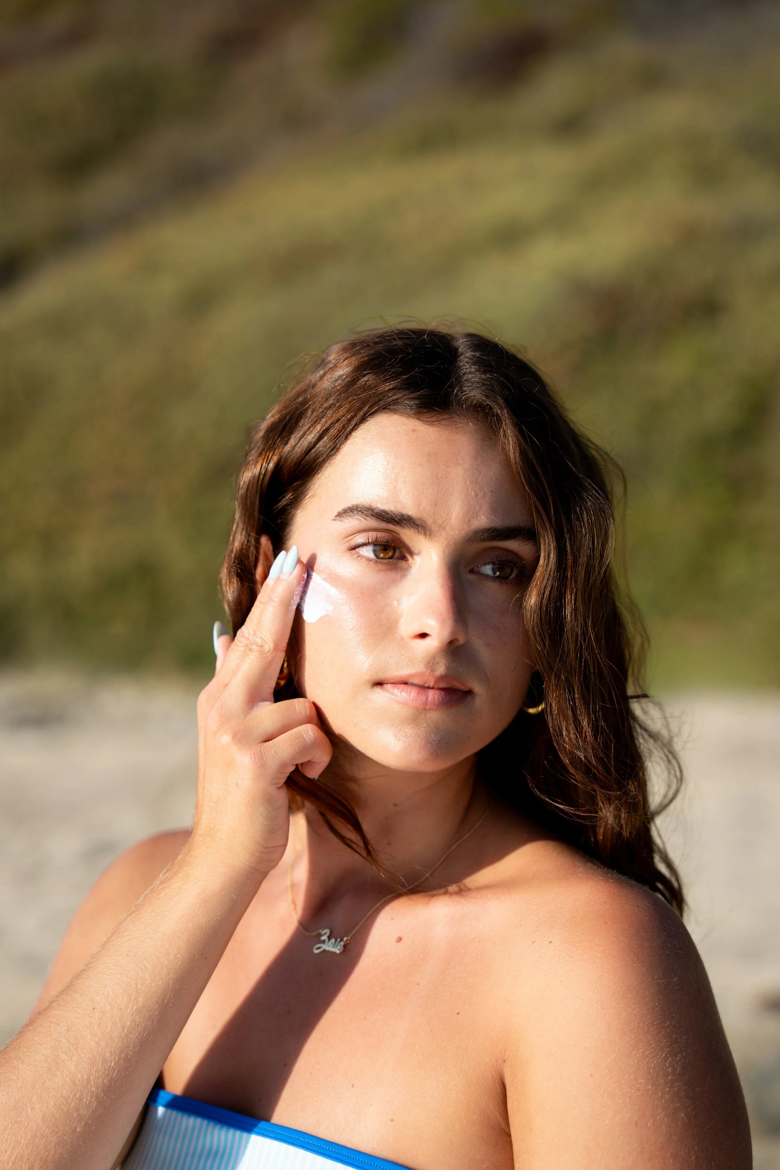 A woman with long brown hair and light brown eyes is outdoors, applying sunscreen to her face. She is wearing a strapless blue and white striped dress and a necklace with the word 'Love.' The background is blurred green nature.