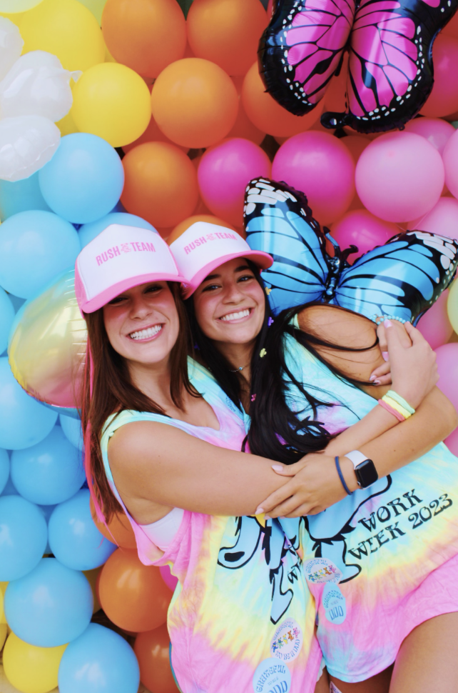 Two women wearing matching tie-dye tank tops and pink caps with white fronts that say 'Rush Team' are embracing and smiling. Behind them is a colorful balloon wall with pink, orange, yellow, blue, and white balloons, and a butterfly-shaped balloon wi