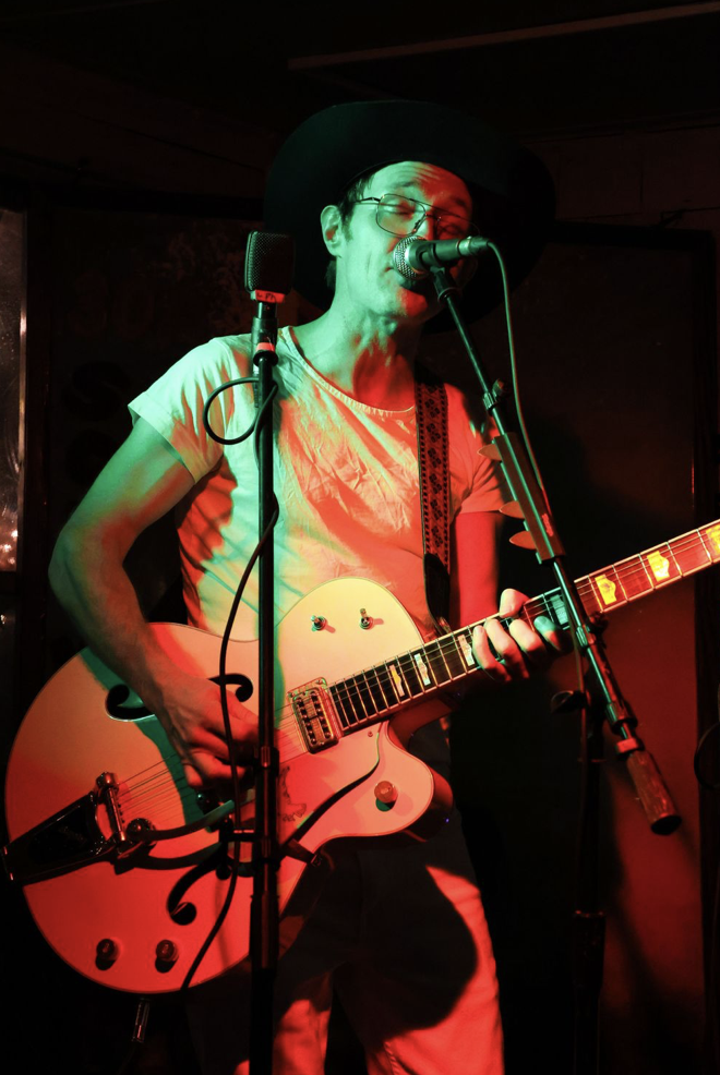 A musician with glasses and a wide-brimmed black hat playing an electric guitar and singing into a microphone, illuminated by red and green stage lighting.