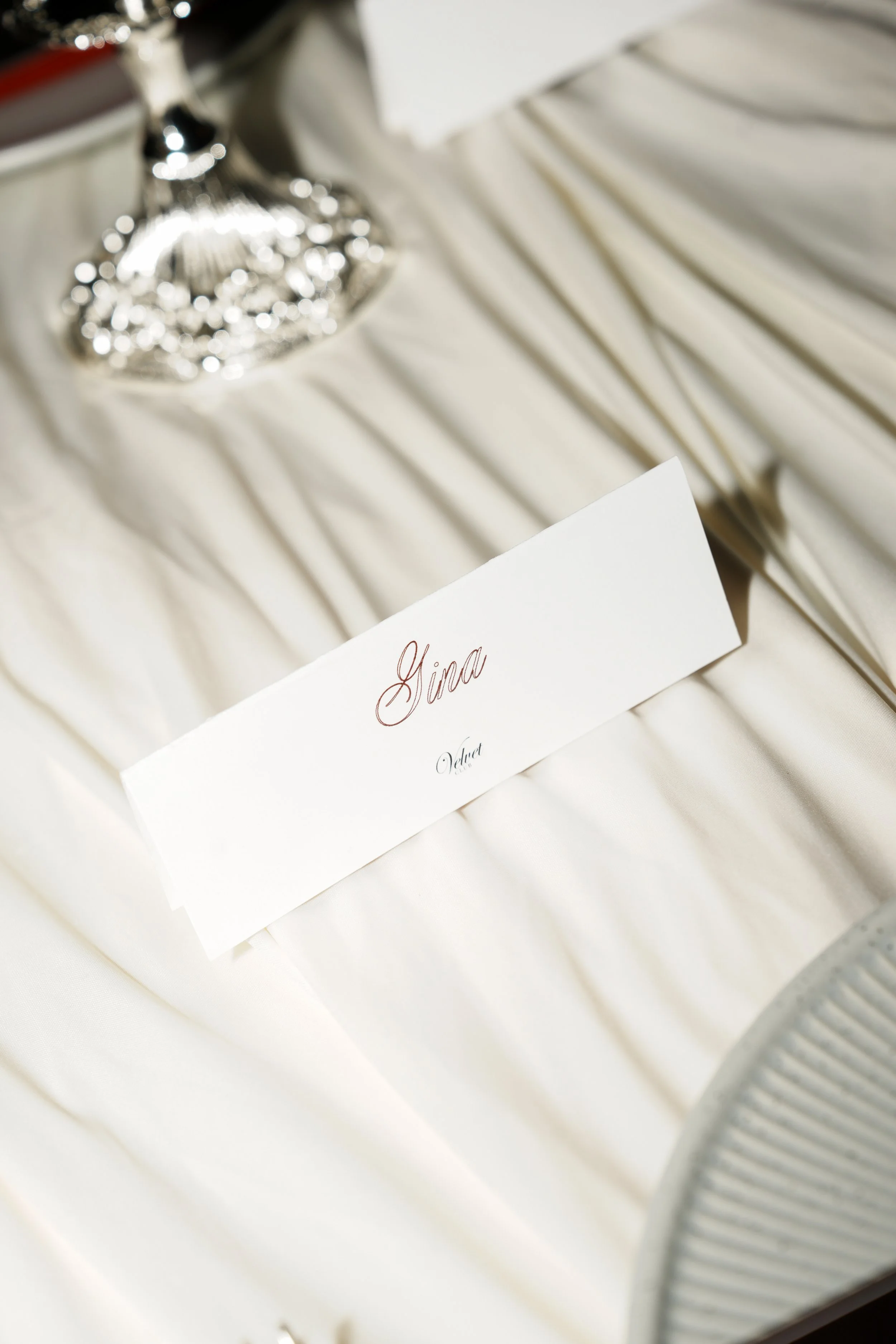 A white table cloth with a place card that says 'Gina' and a silver utensil. In the background, a decorative silver floral item and a white napkin fold are visible.