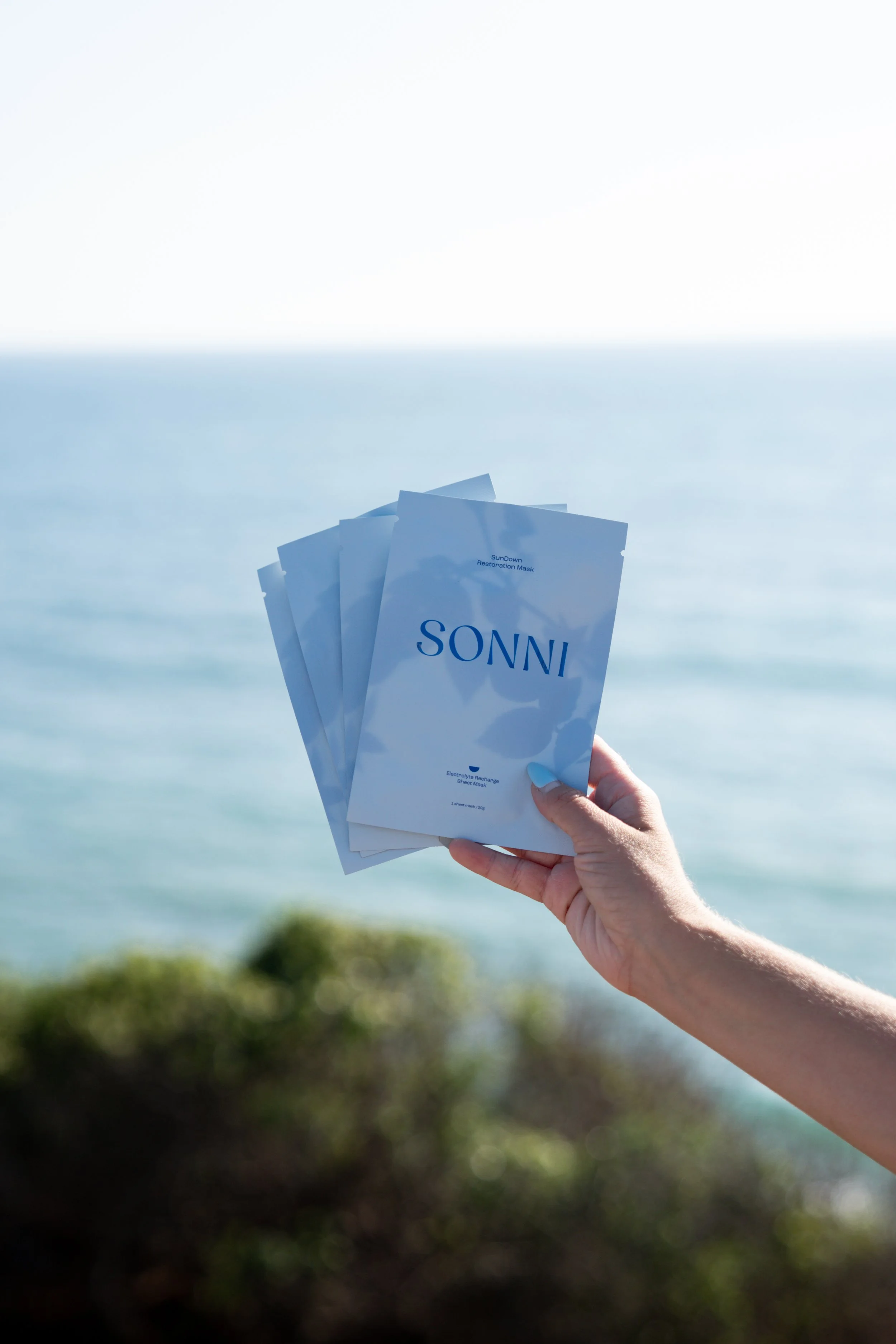 Person holding several blue and white packages labeled 'SunDown Restoration Mask' near a body of water and greenery.