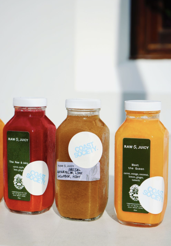 Three jars of colorful juice with Green Coast Society labels, each with white lids. The left jar is red, the middle jar is orange, and the right jar is bright yellow.