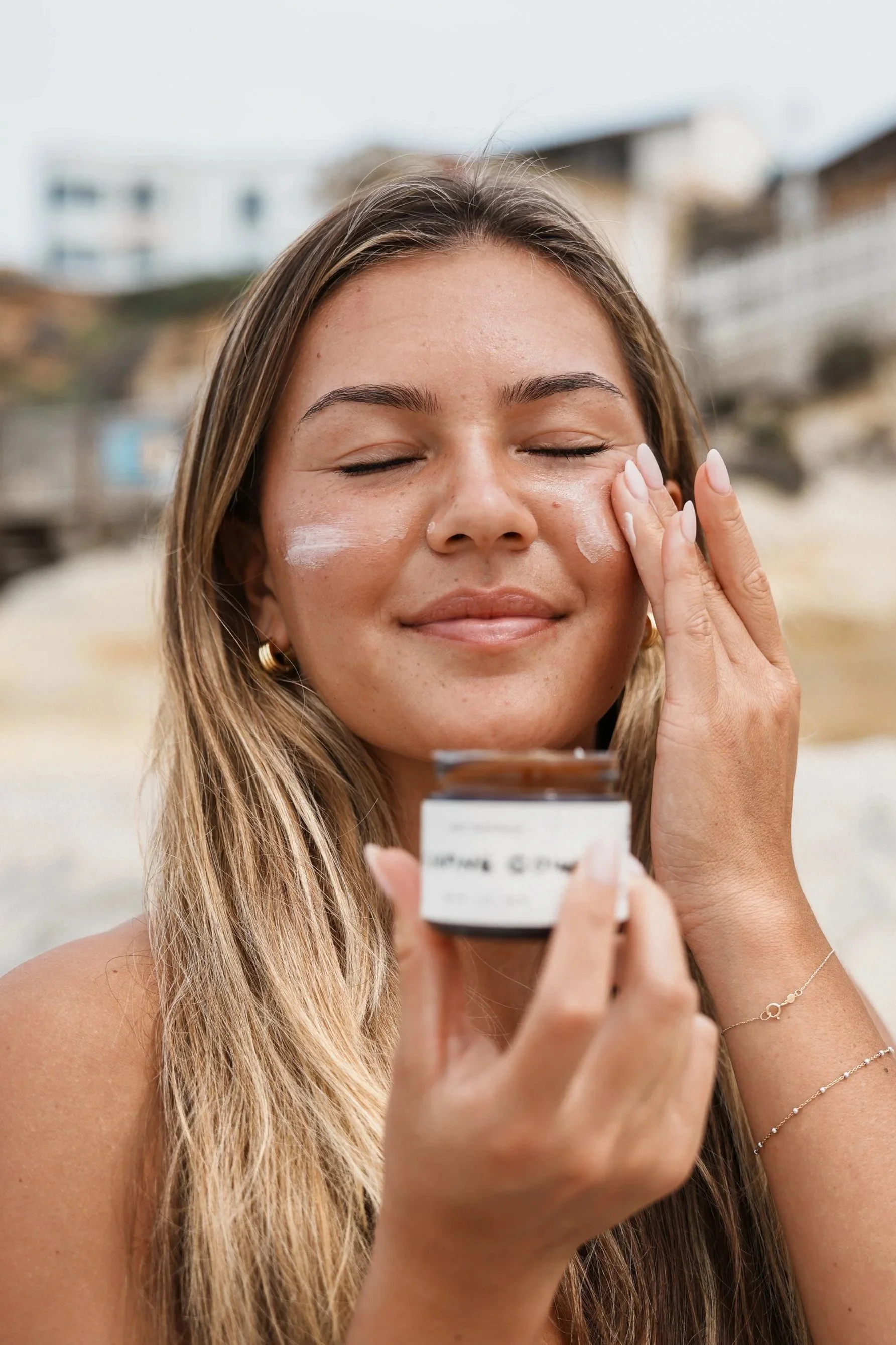 A woman with long blonde hair, closed eyes, and a gentle smile applying lotion to her face at the beach, holding a small jar of skincare product.