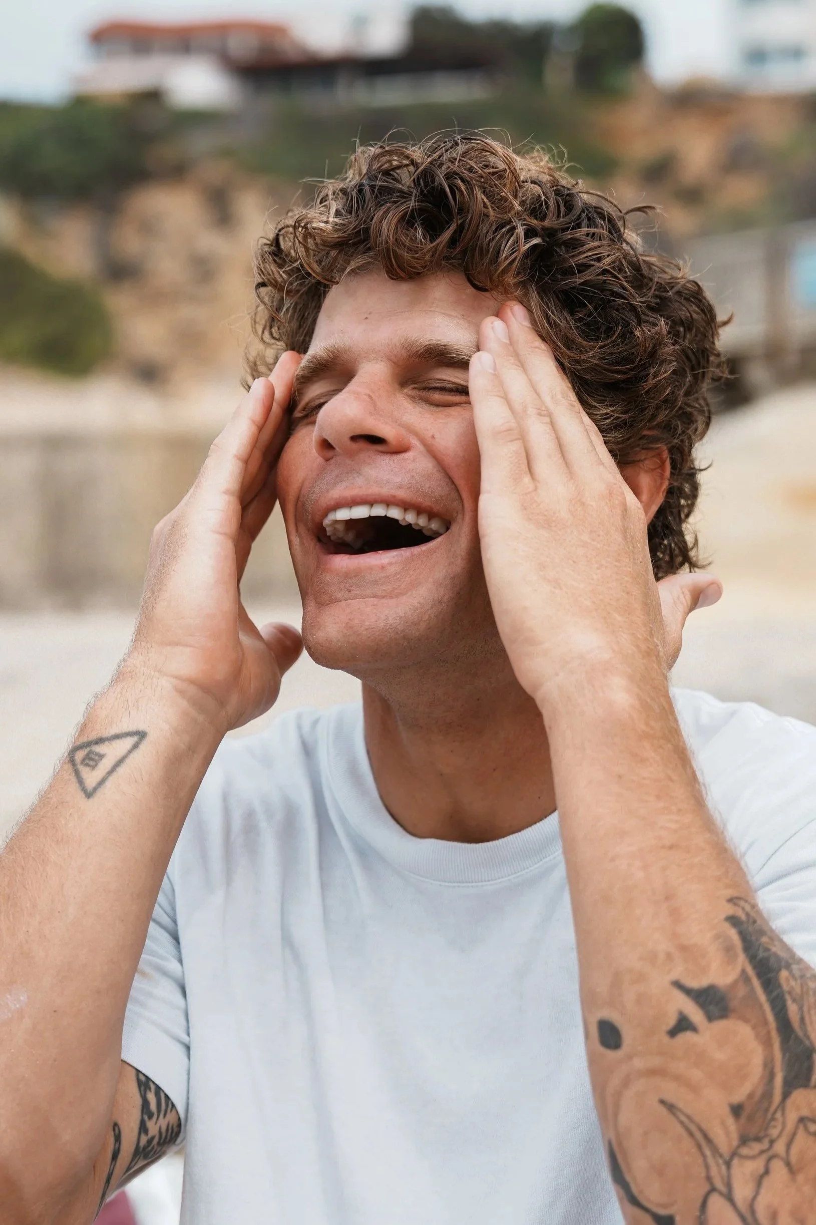 A man with curly brown hair, tattoos on both arms, and wearing a white shirt, appears to be at the beach with his eyes closed and hands on his temples, smiling or laughing. The background features sandy terrain, blurred houses, and cloudy skies.