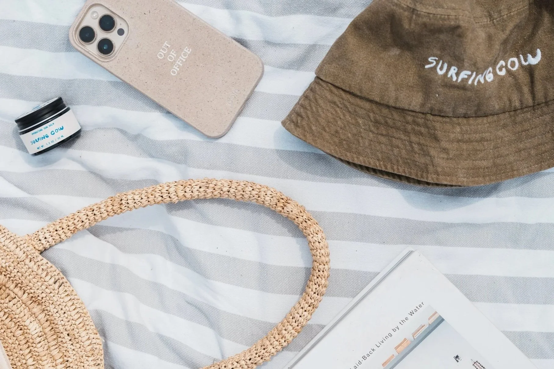 A flat lay of items on a striped fabric surface, including a straw bag, a brown cap, a smartphone, a small bottle, a tan note card, and part of a person's leg wearing shorts.