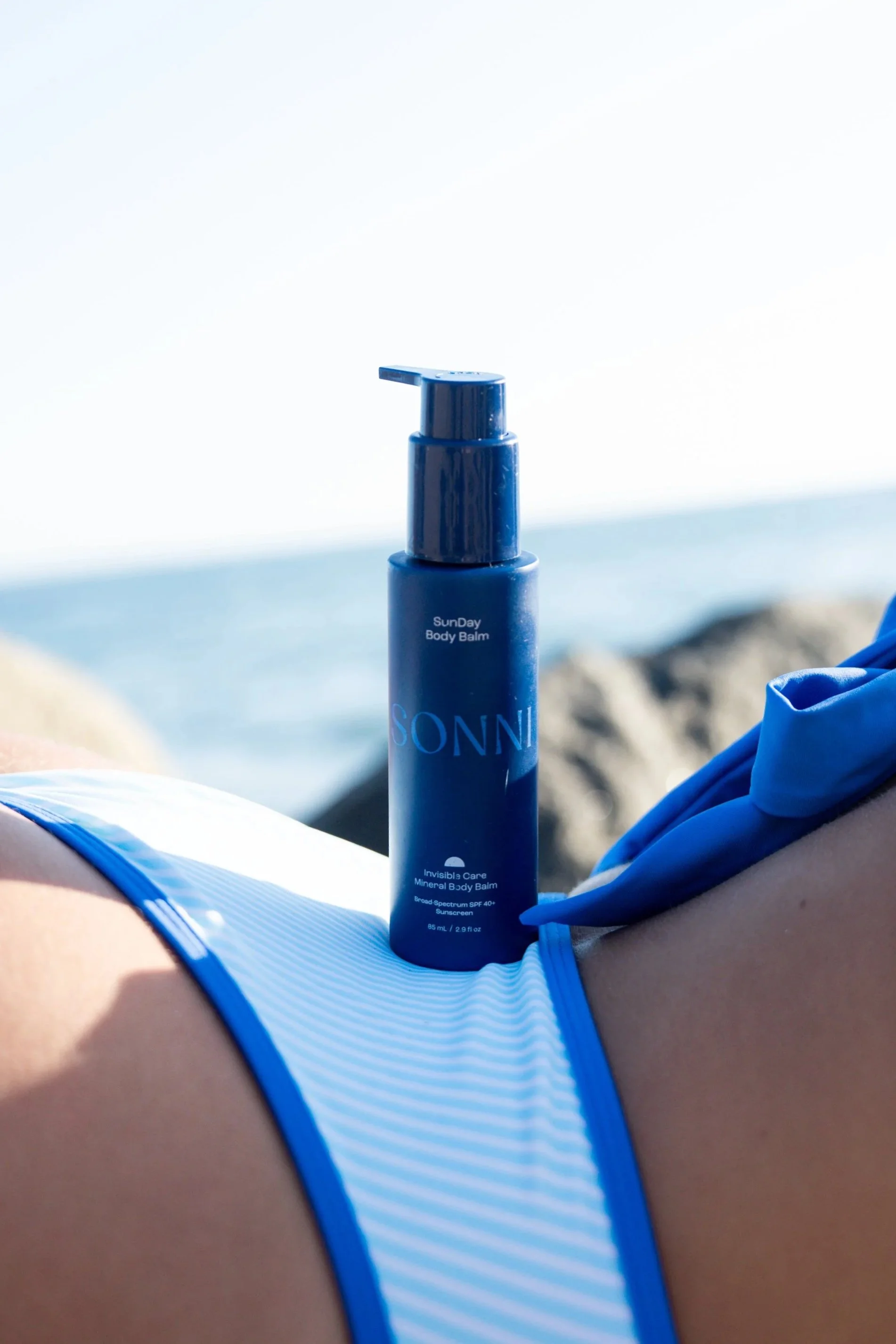 A person lying on a beach wearing a blue and white swimsuit with a blue bottle of SunDay Body Balm resting on their chest, with blurred rocky shoreline and ocean in the background.