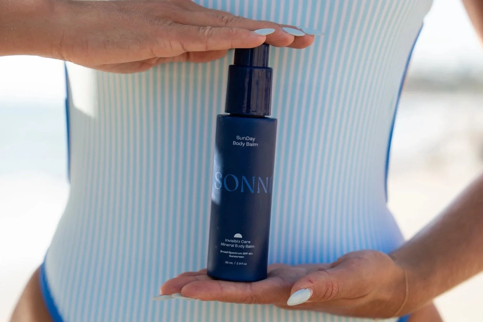 Close-up of a woman wearing a striped swimsuit, holding a bottle of sunscreen lotion on her stomach with one hand while her other hand holds the sunblock on her stomach.