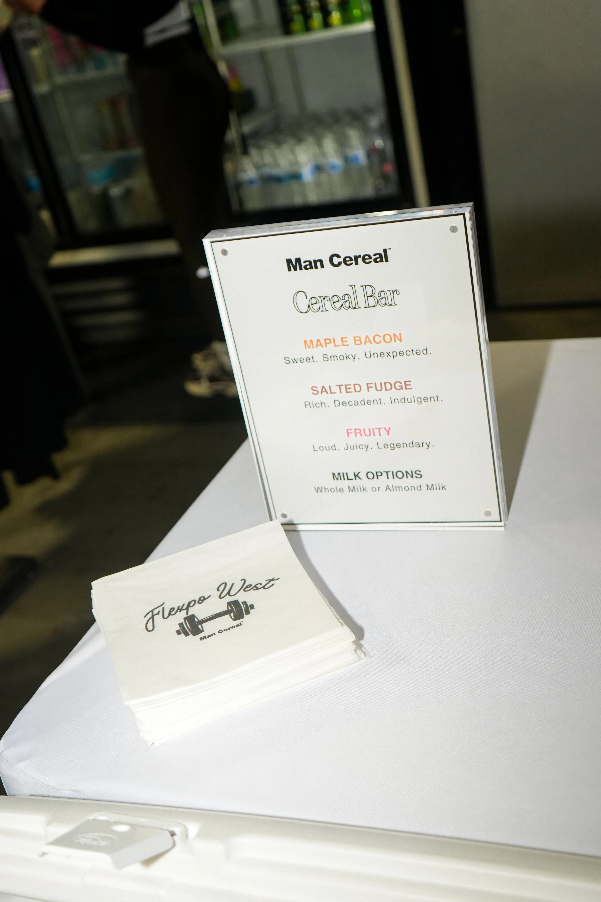A menu sign for 'Man Cereal' cereal bar featuring flavor options like Maple Bacon, Salted Fudge, and Fruity, with descriptions. Next to the sign are stacked paper napkins with a logo for 'Fleppo West.'