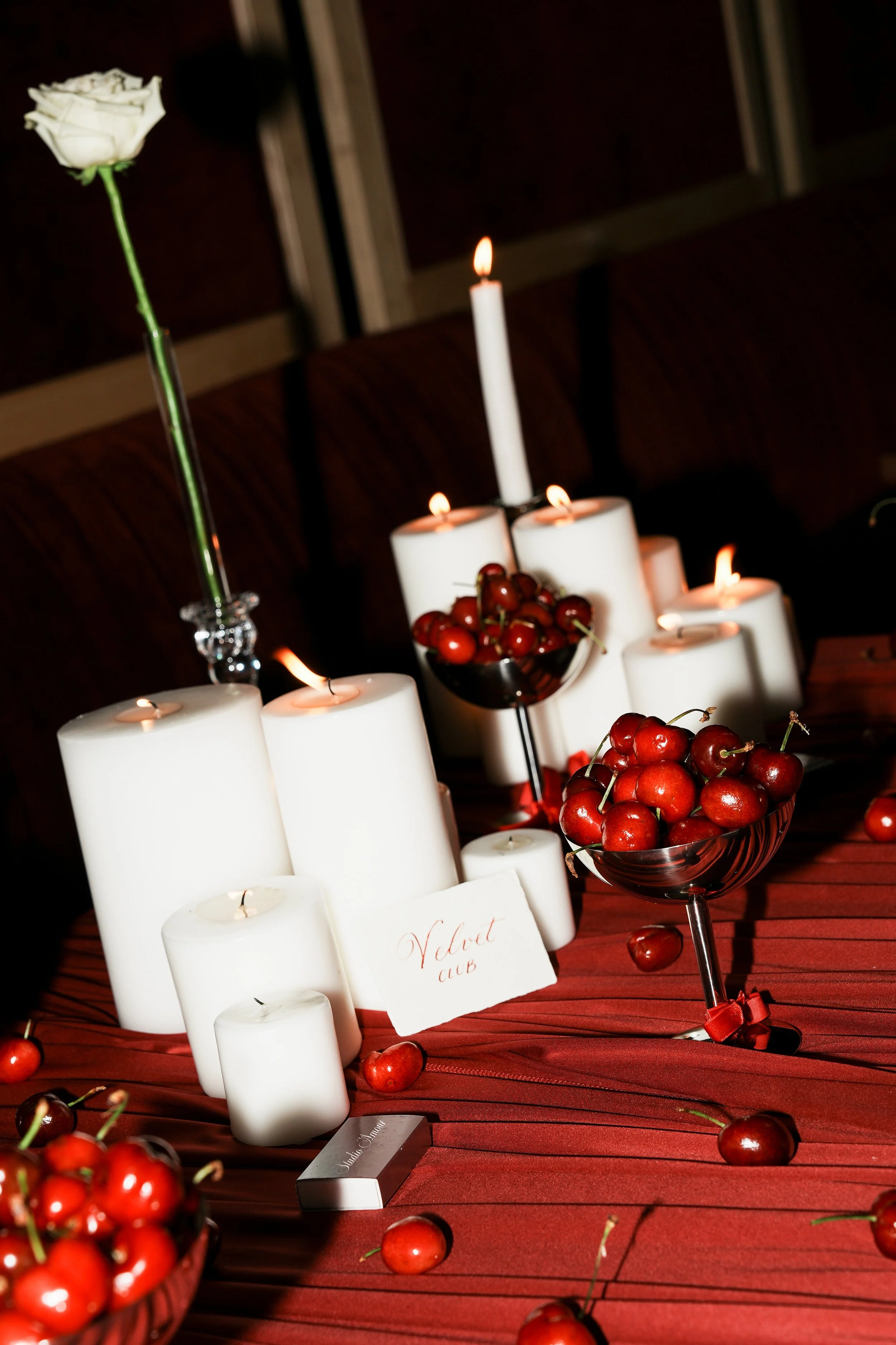 A table decorated with white candles, a bowl of cherries, and a white rose in a tall glass.