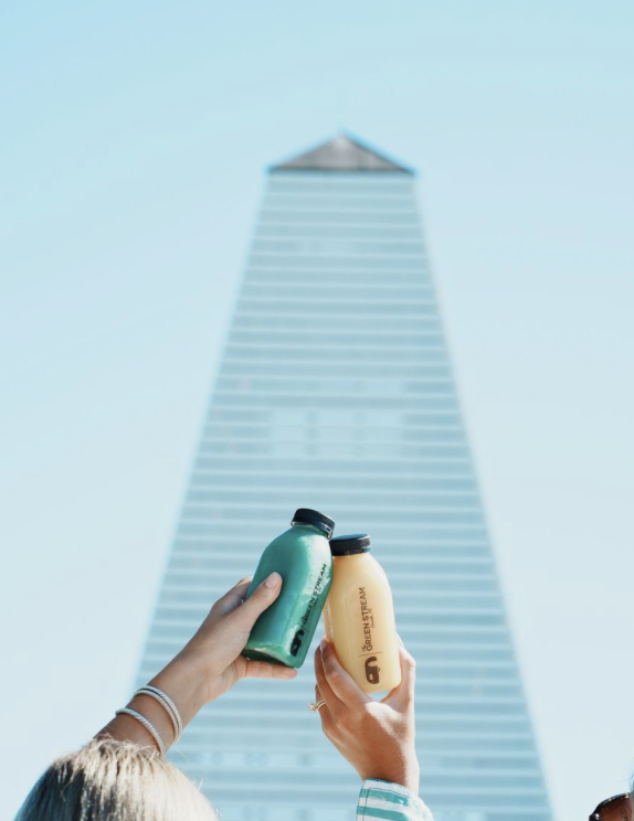 Two hands holding bottles of drink with a tall modern building in the background.