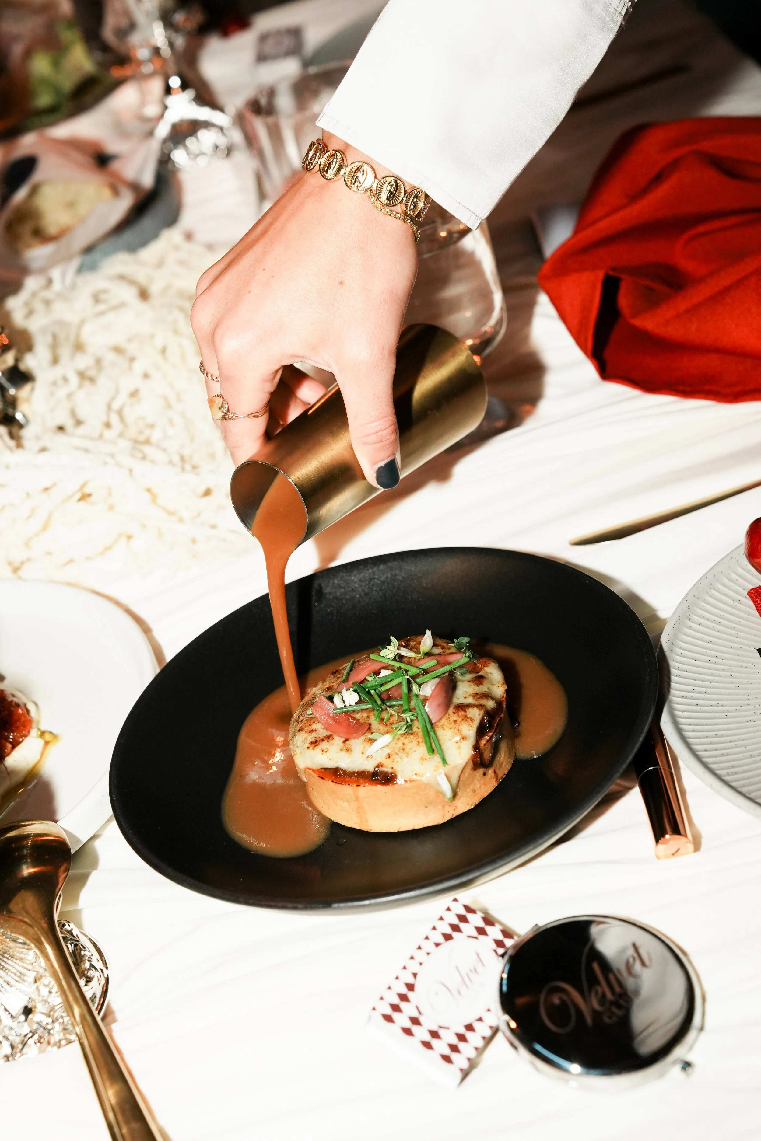 A hand with gold jewelry is pouring gravy over a baked dish topped with vegetables and herbs on a black plate. The table has various dishes, silverware, and glasses.