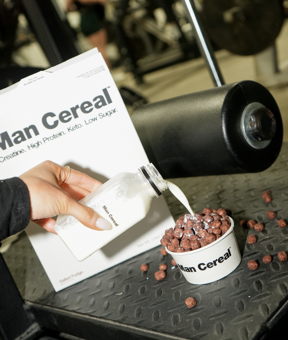 Person pouring milk into a bowl of chocolate cereal at a fitness event, with a Man Cereal box and a foam roller in the background.
