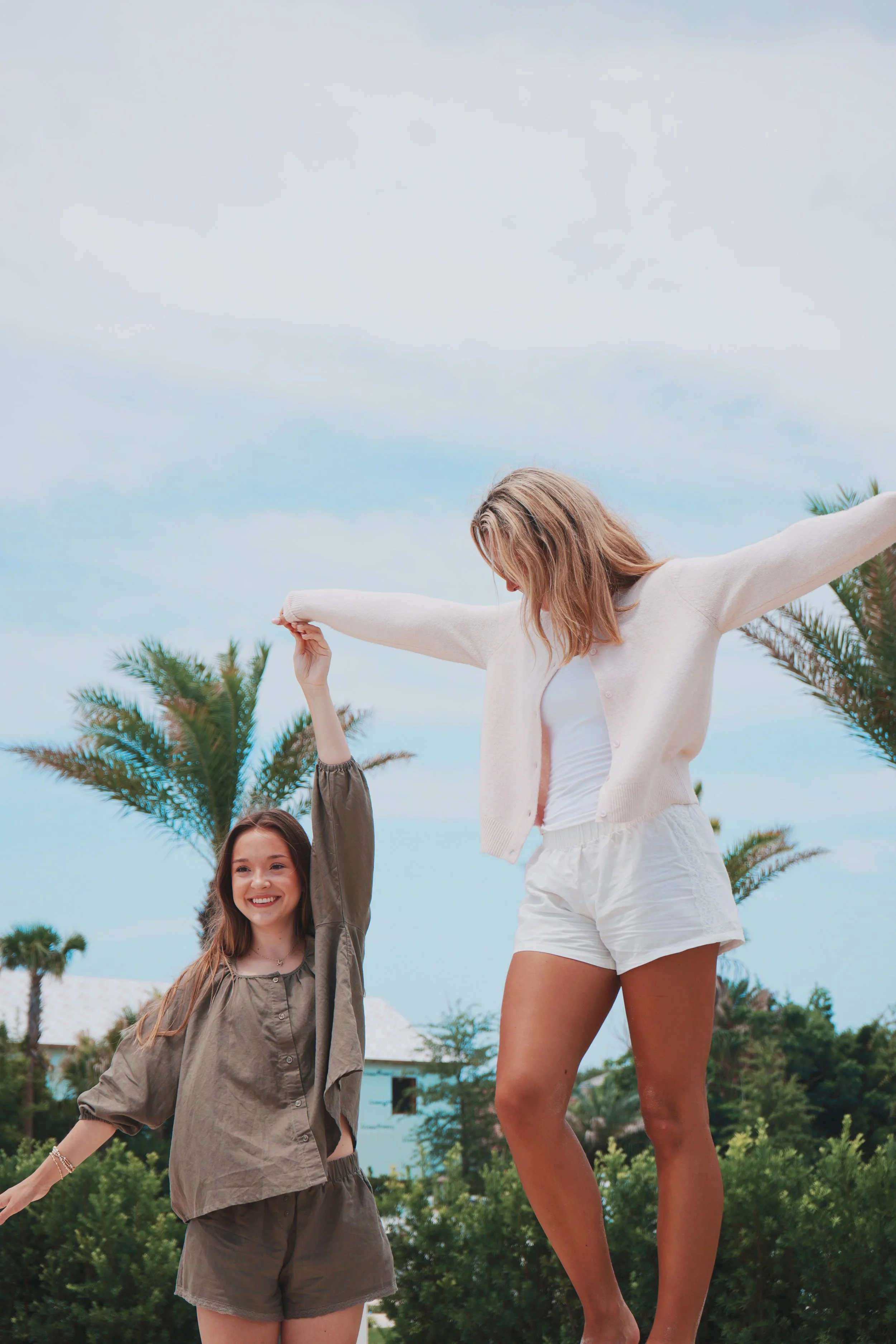 Two women are outdoors, one smaller and one taller, holding hands in the air. The taller woman is balancing on her toes with arms outstretched, and the smaller woman is smiling at the camera, wearing a loose jacket. Palm trees and a blue sky are in t