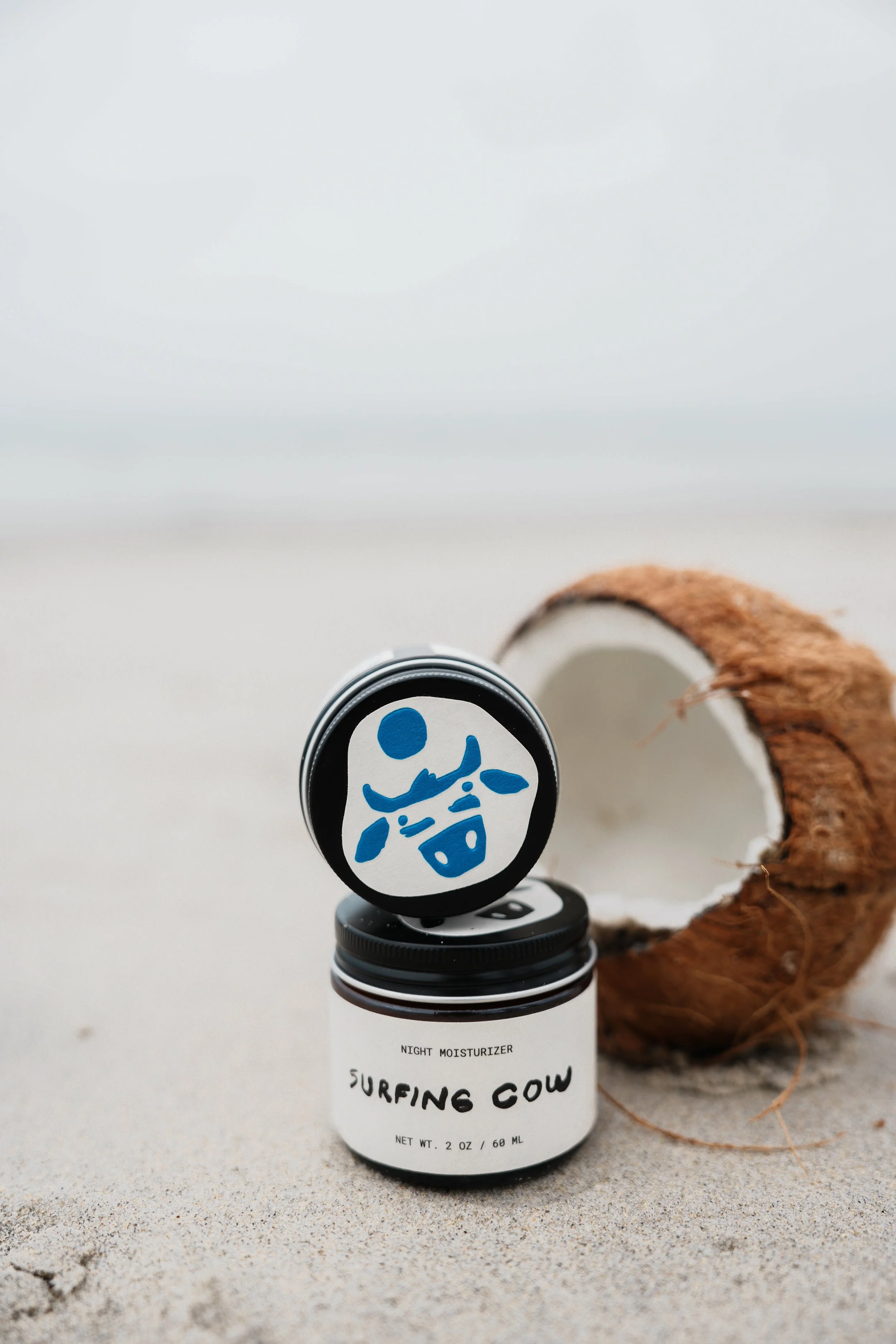 A small white bottle labeled "Surfing Cow" night moisturizer, placed on a surface with sand, next to a brown, fuzzy, circular object that resembles a coconut husk.