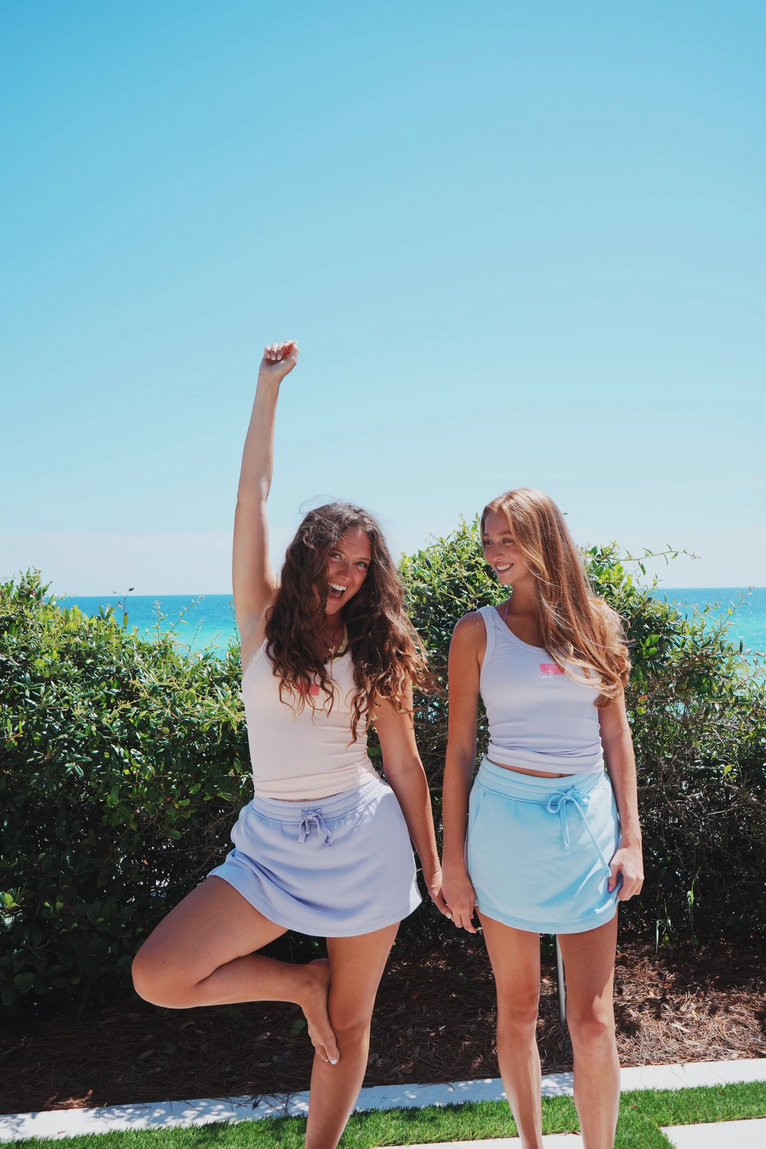 Two young women in beachwear standing outside near green bushes, with the ocean and clear blue sky in the background.