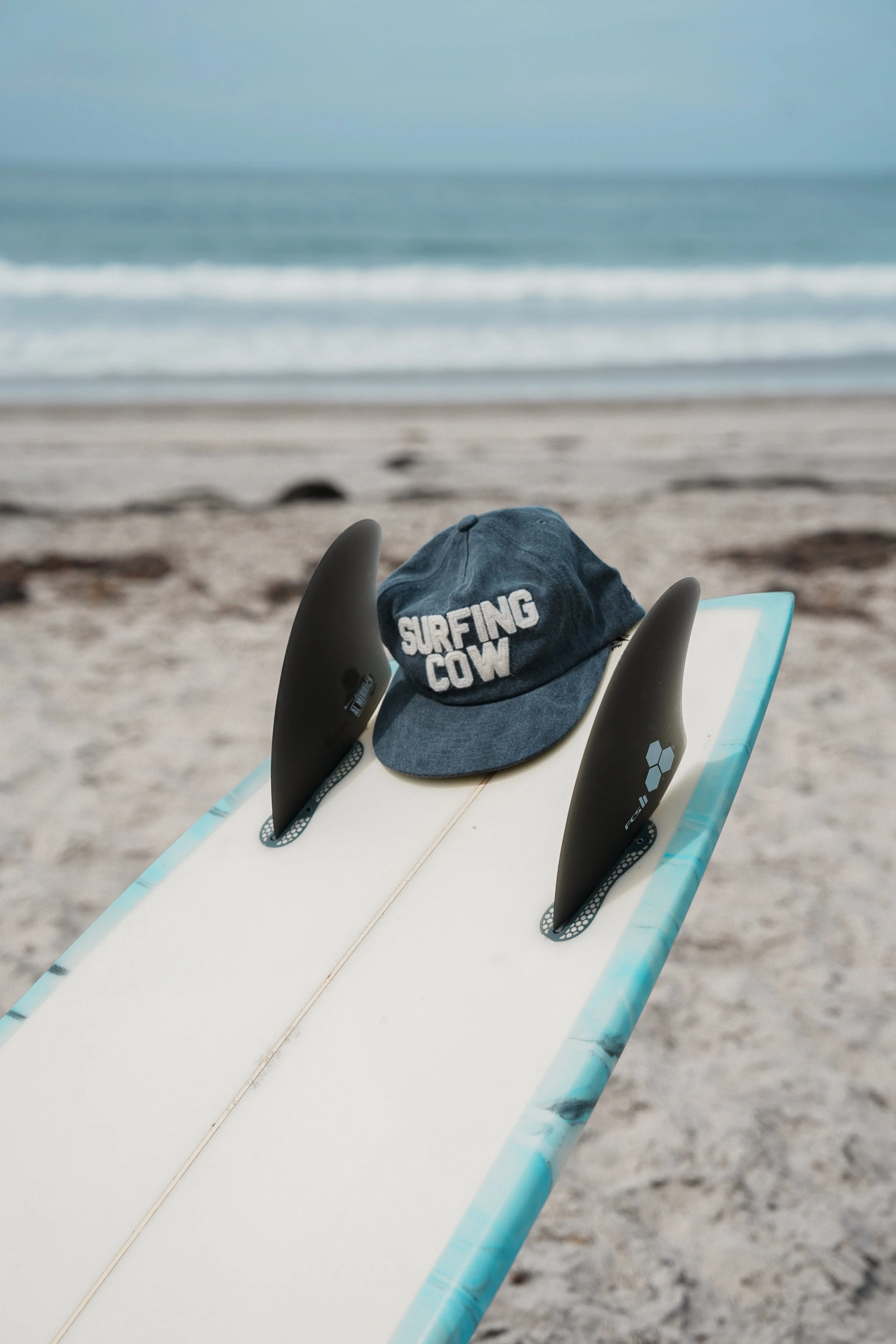 A surfboard on the sandy beach with a surfboard leash attached, a blue cap that says "SURFING COW", and a blue and white surfboard in the foreground, with the ocean waves and sky in the background.
