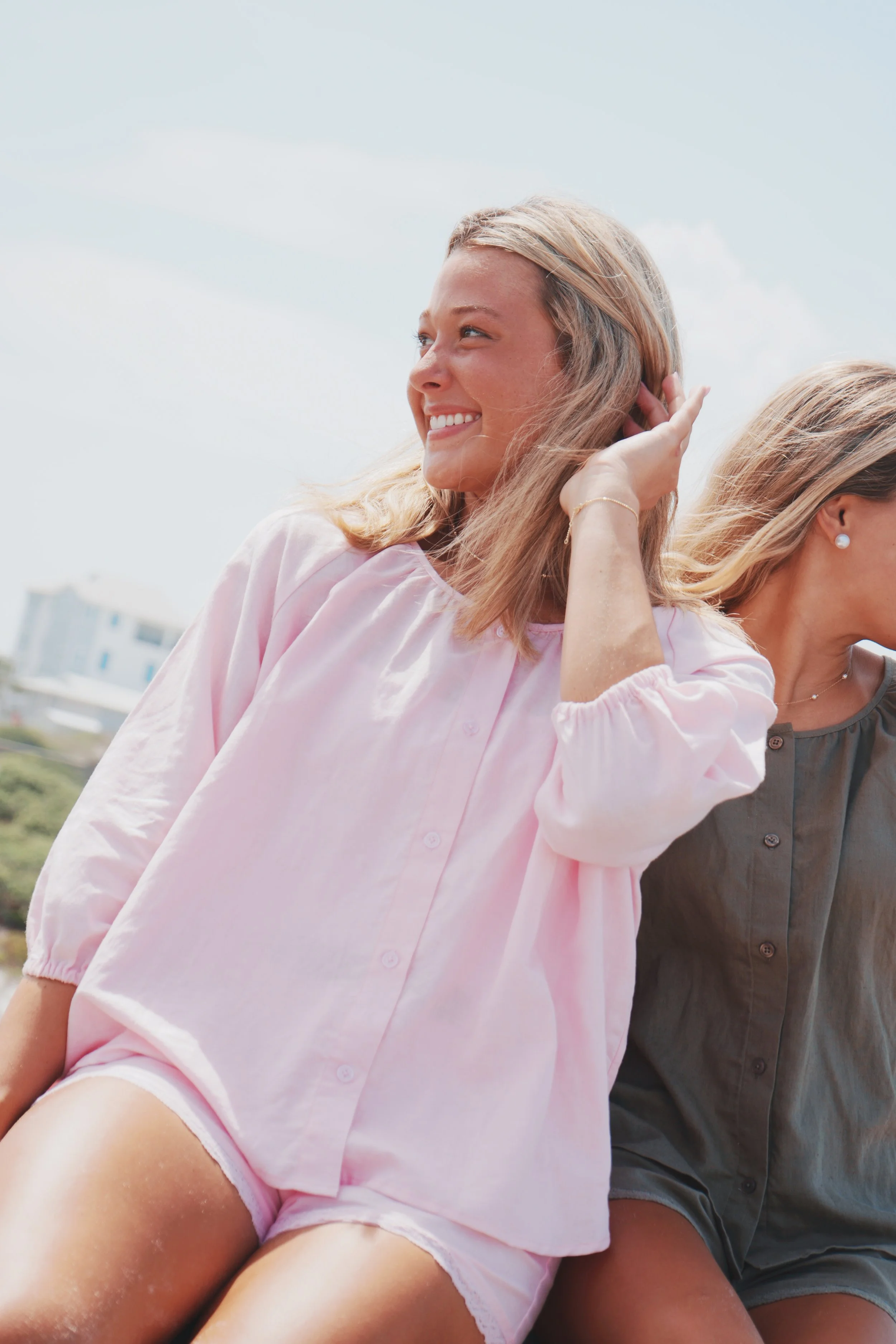 A woman with blonde hair smiling and adjusting her hair outdoors on a sunny day, wearing a light pink shirt. Part of another woman is visible beside her, wearing a dark grey shirt and pearl earrings.